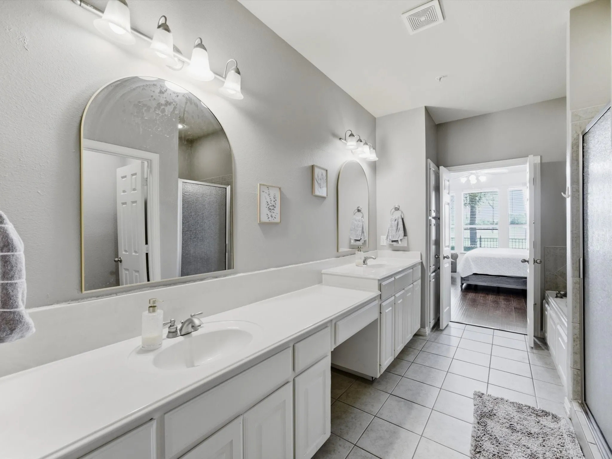 Bathroom featuring dual vanities, arched mirrors, a built-in makeup counter, tiled flooring, and a shower enclosure