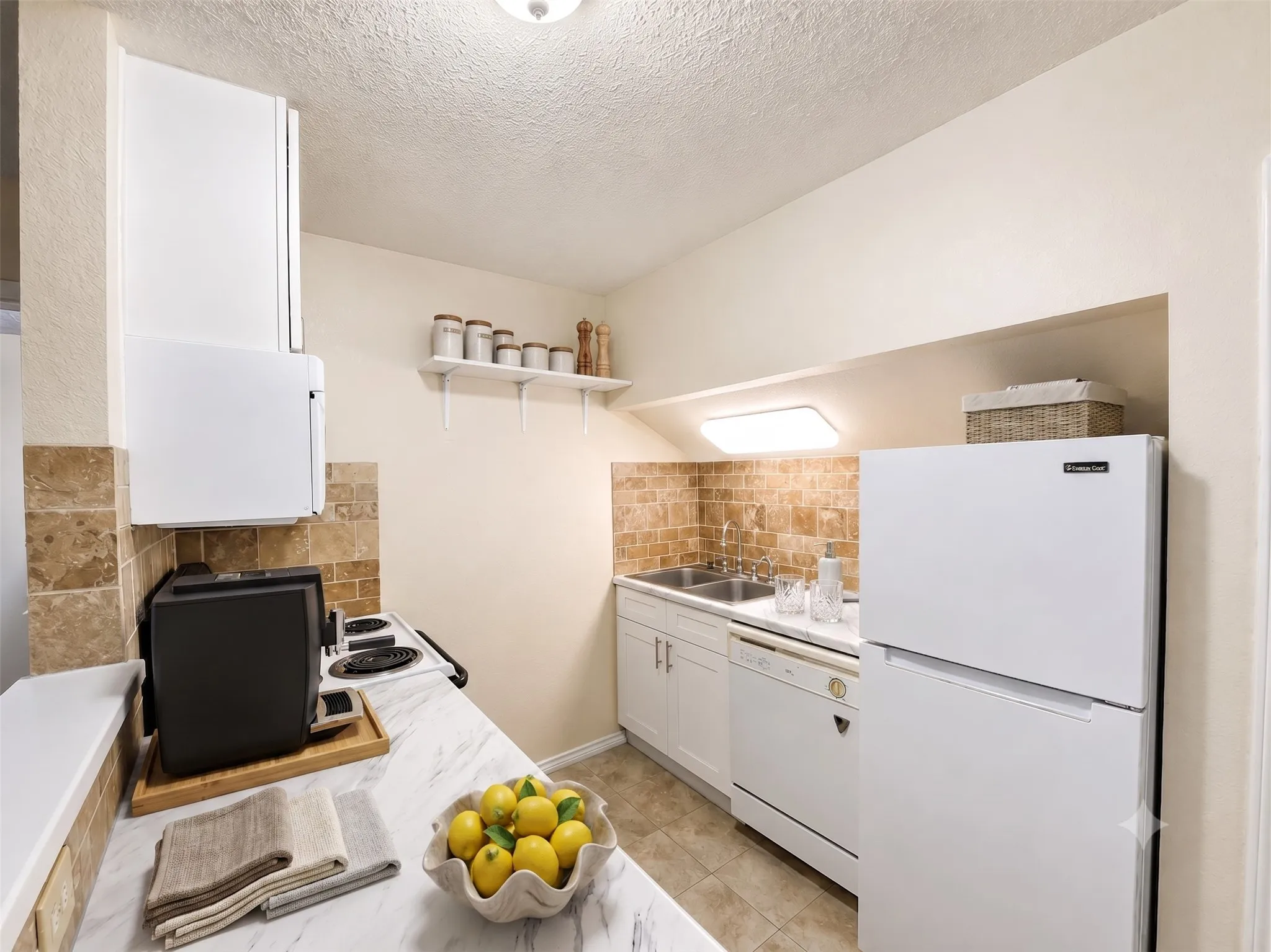 Compact kitchen featuring a double basin sink, white cabinetry, integrated dishwasher, and a refrigerator-Virtually Staged