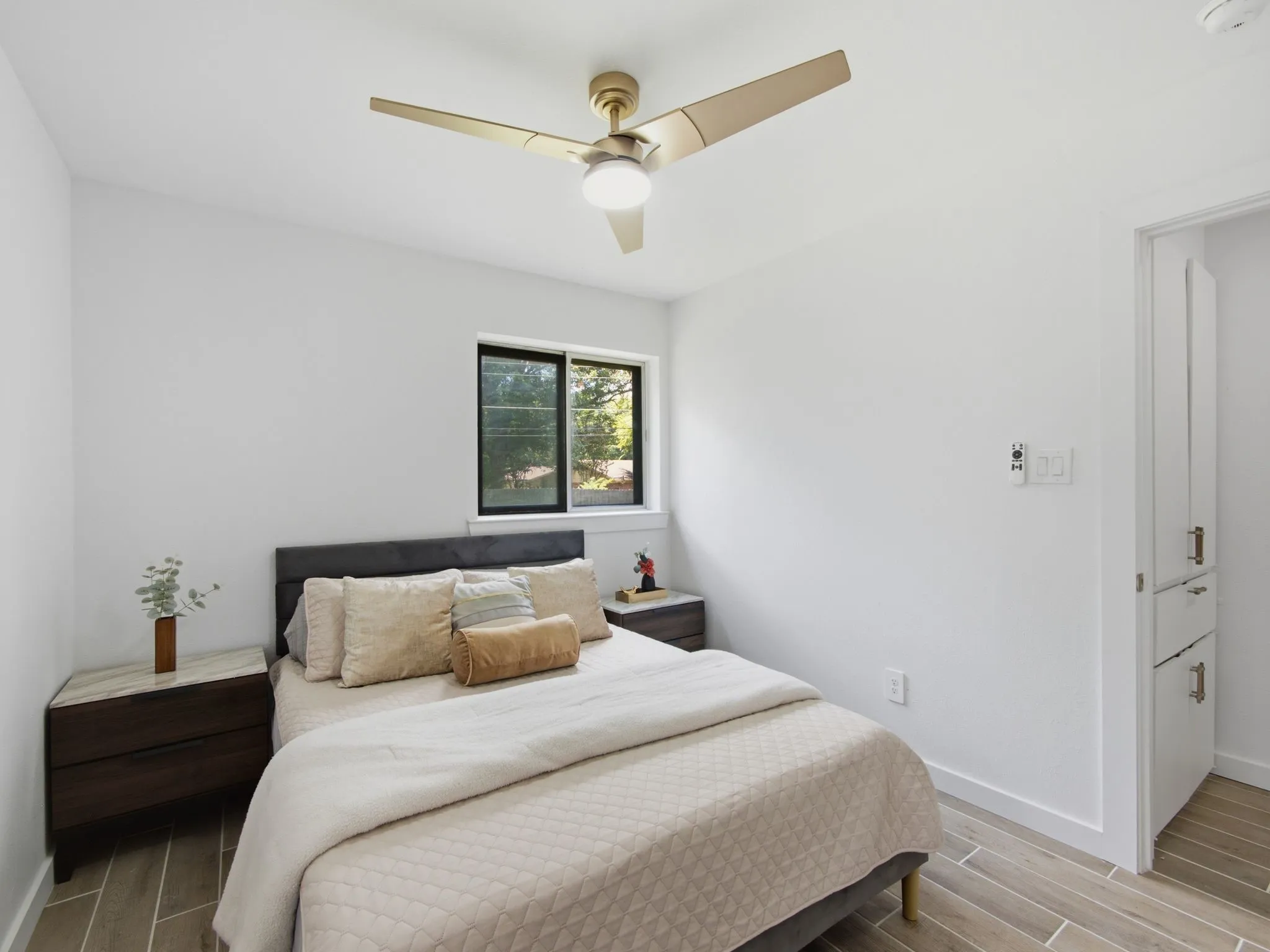 Gold-tone ceiling fan with integrated lighting, wood-finish flooring, and a single window with dark framing