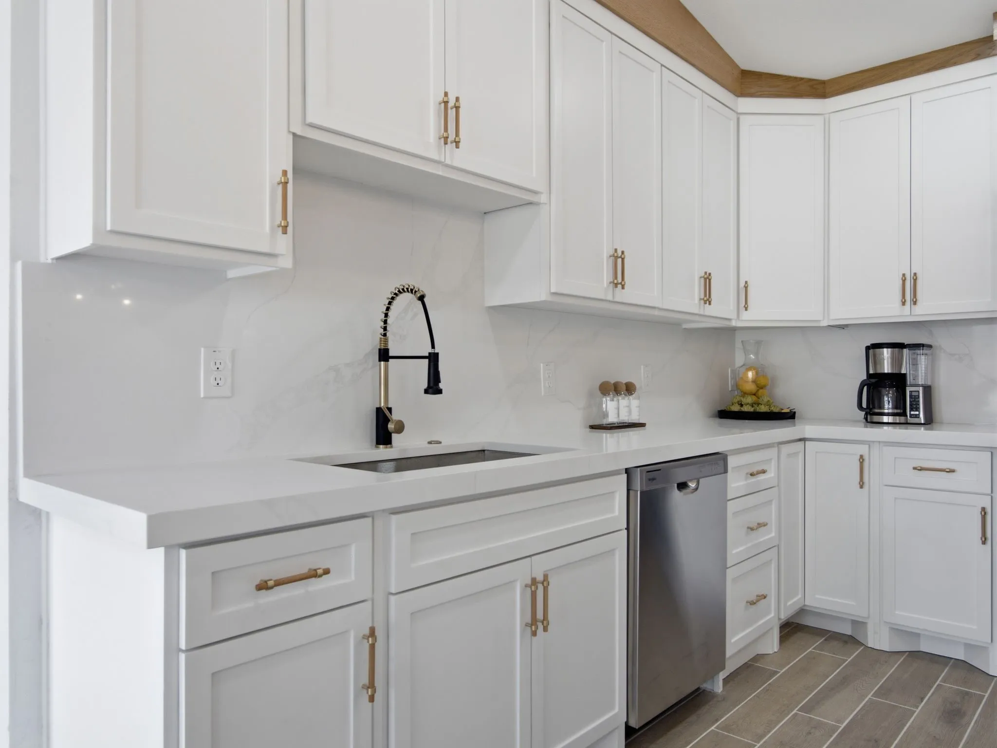 White shaker cabinetry with gold-toned hardware, solid surface countertops, a stainless steel sink with a black and gold-toned faucet, and a tile backsplash