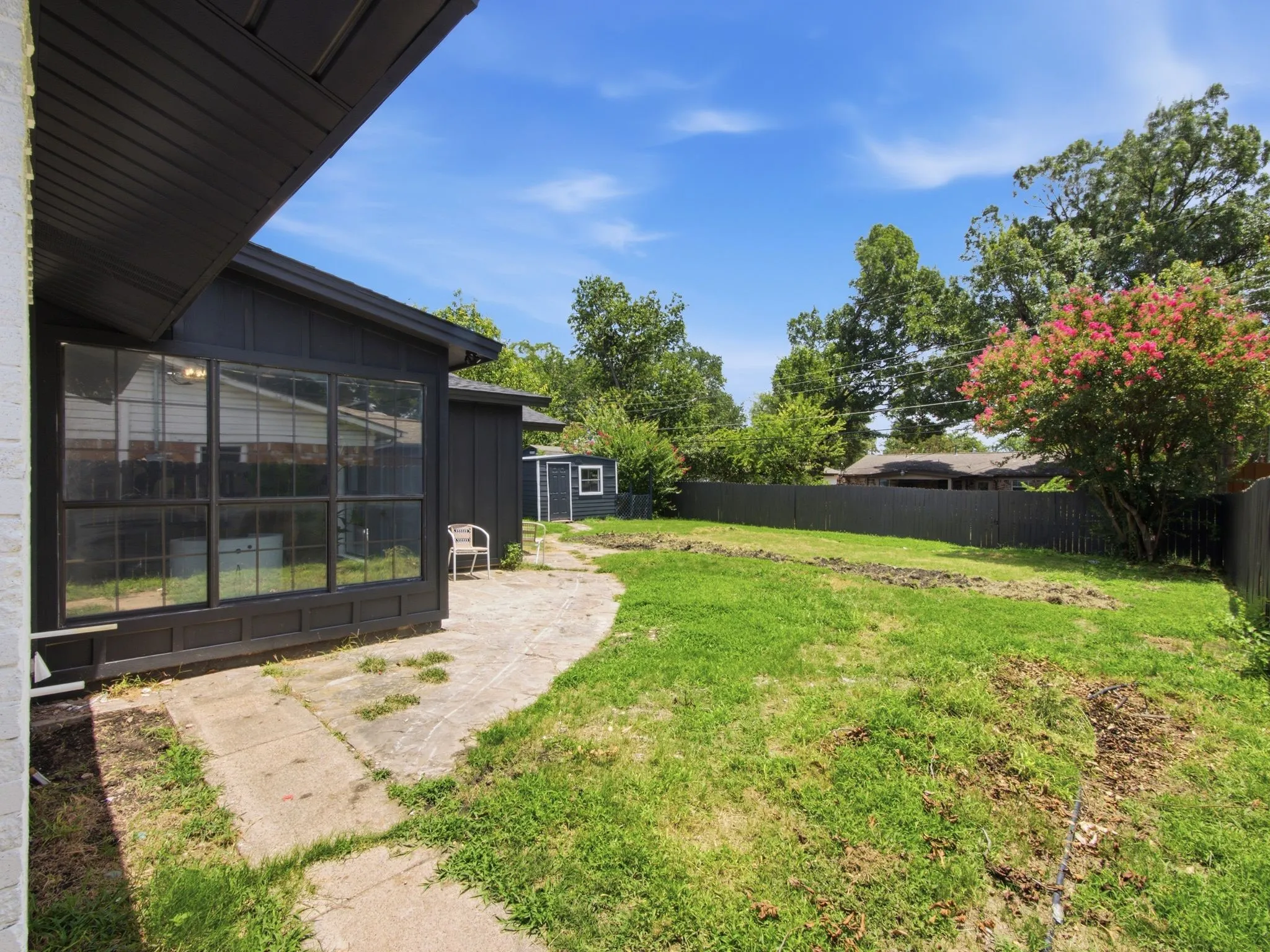 Spacious backyard featuring a concrete patio, a sunroom with large windows, a detached shed, a privacy fence, and mature trees