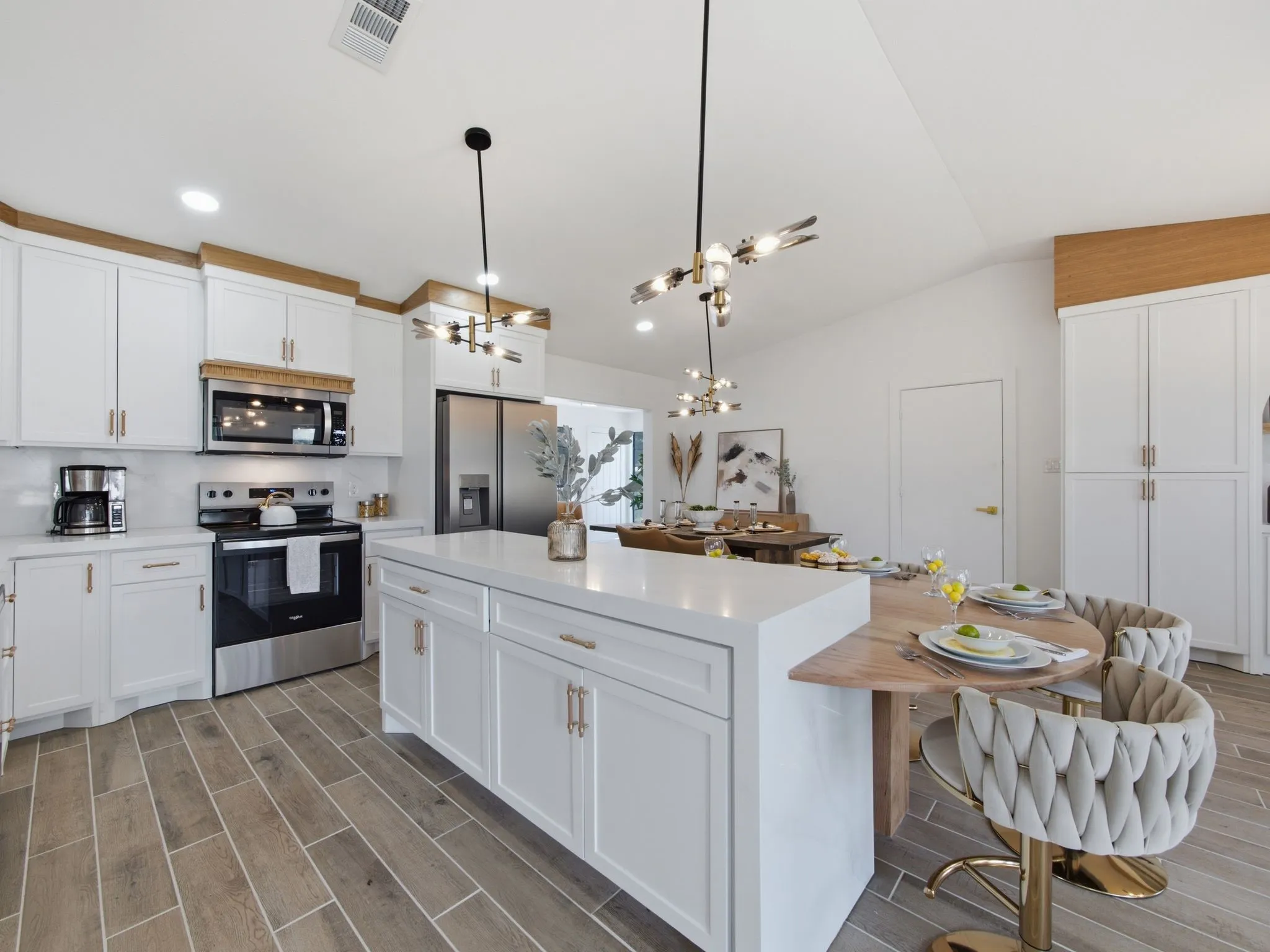 White shaker cabinetry with gold-tone hardware, stainless steel appliances, wood-finish tile flooring, a large kitchen island with a white countertop, and contemporary pendant lighting fixtures