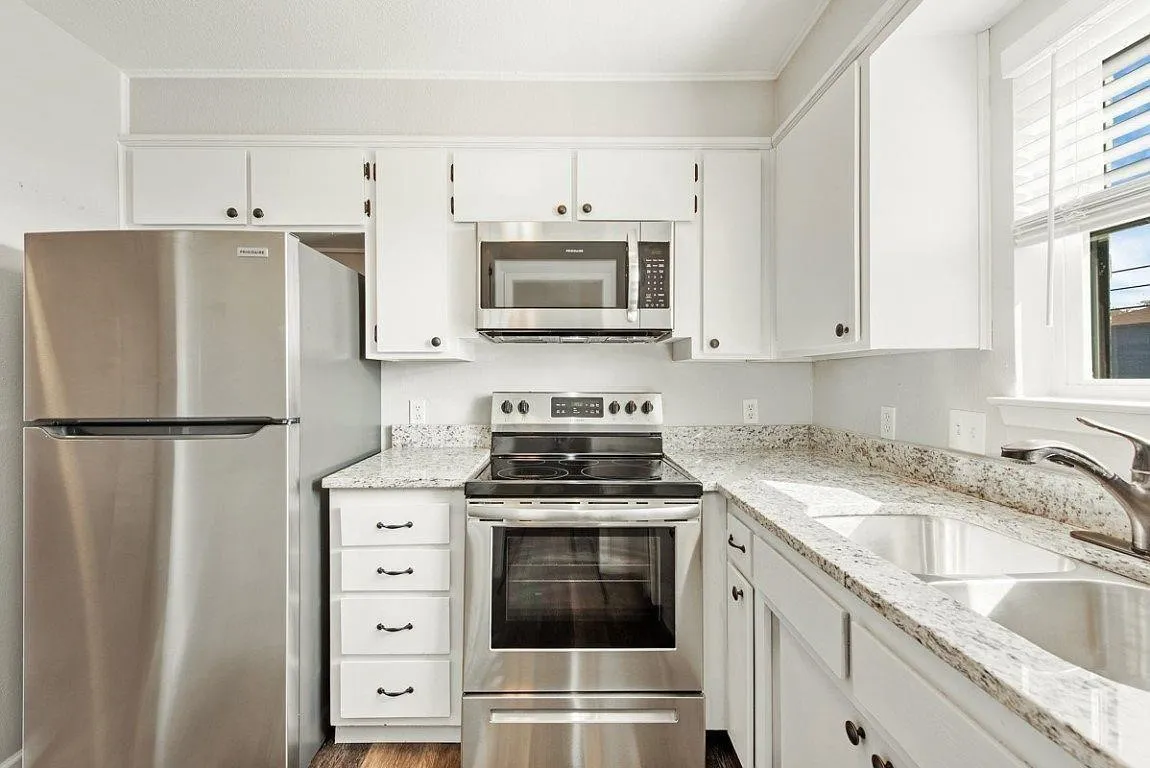 Kitchen featuring white cabinetry, stainless steel appliances, speckled countertops, a double basin sink, and a window with blinds