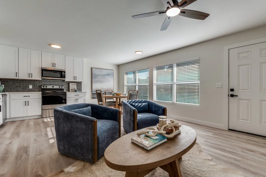 Open-concept living space featuring wood-finish flooring, a contemporary ceiling fan, multiple windows with blinds, white kitchen cabinetry with stainless steel appliances, and a dining area