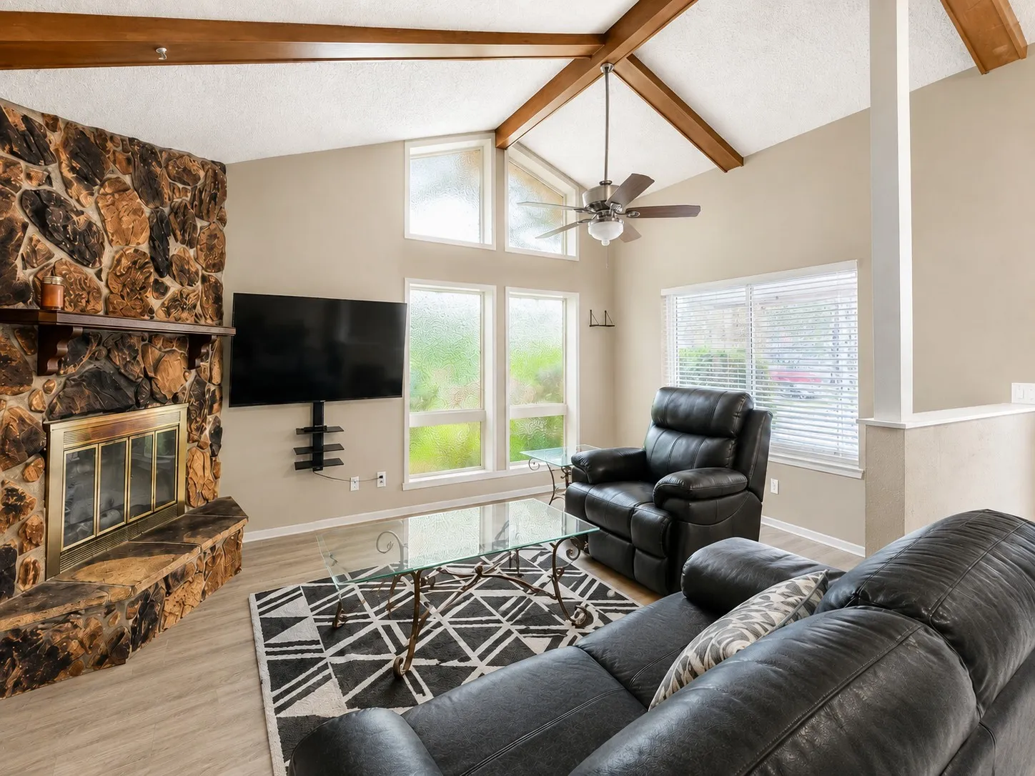Vaulted ceilings with exposed wood beams, stone-finish fireplace with a wood mantle, large windows, and wood-finish flooring