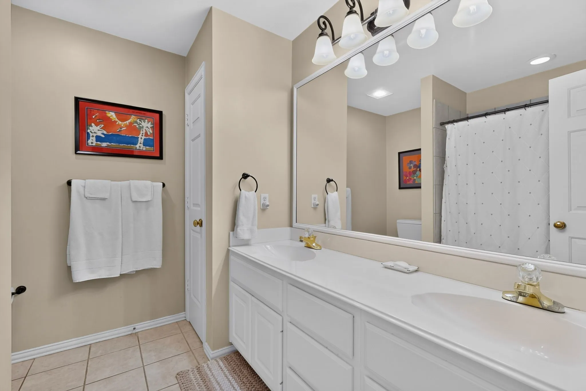 Double vanity with integrated sinks, large mirrored wall, multi-bulb vanity lighting, and tiled flooring