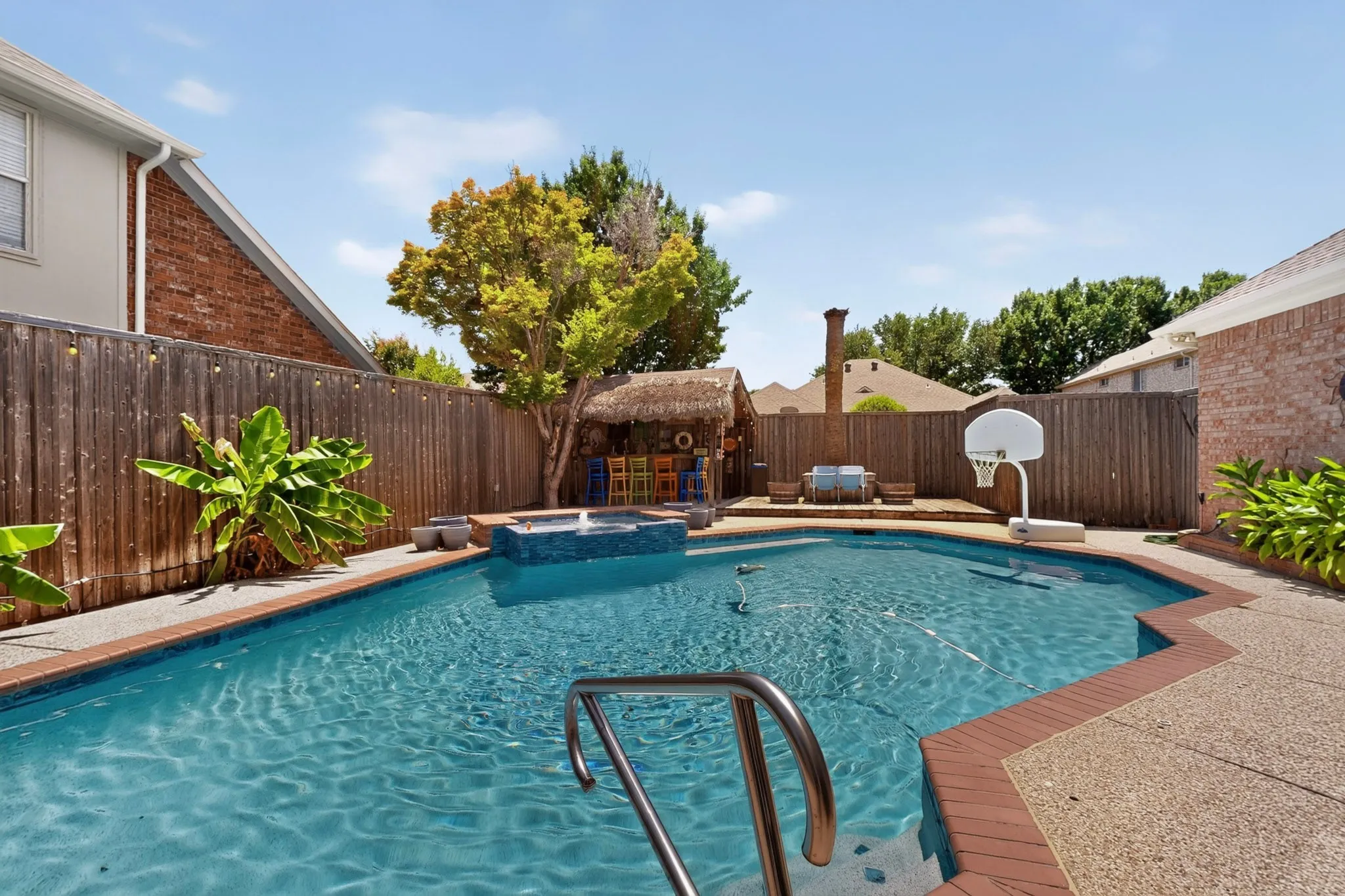 Resort-style backyard oasis featuring a sparkling in-ground swimming pool with integrated spa, brick decking, mature landscaping with tropical plantings, wooden privacy fencing, and a basketball hoop for recreational enjoyment