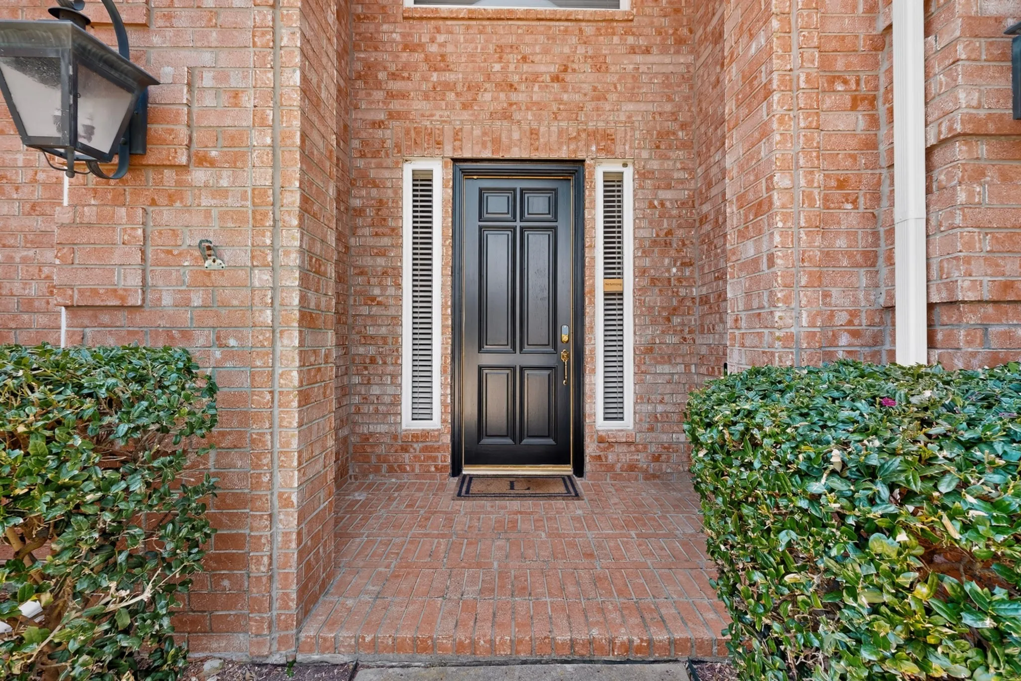 Elegant brick courtyard entry featuring a six-panel black lacquered door with sidelights, complemented by manicured hedge landscaping and traditional lantern sconce lighting