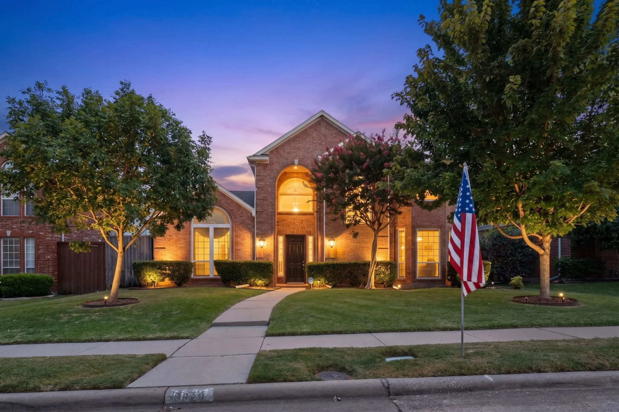 Brick exterior with arched entryway, mature trees, manicured lawn, and exterior uplighting