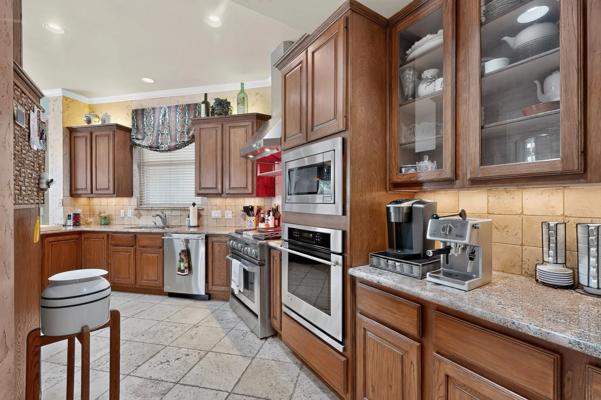 Kitchen featuring custom wood cabinetry, stainless steel appliances, tile flooring, stone countertops, and a tile backsplash