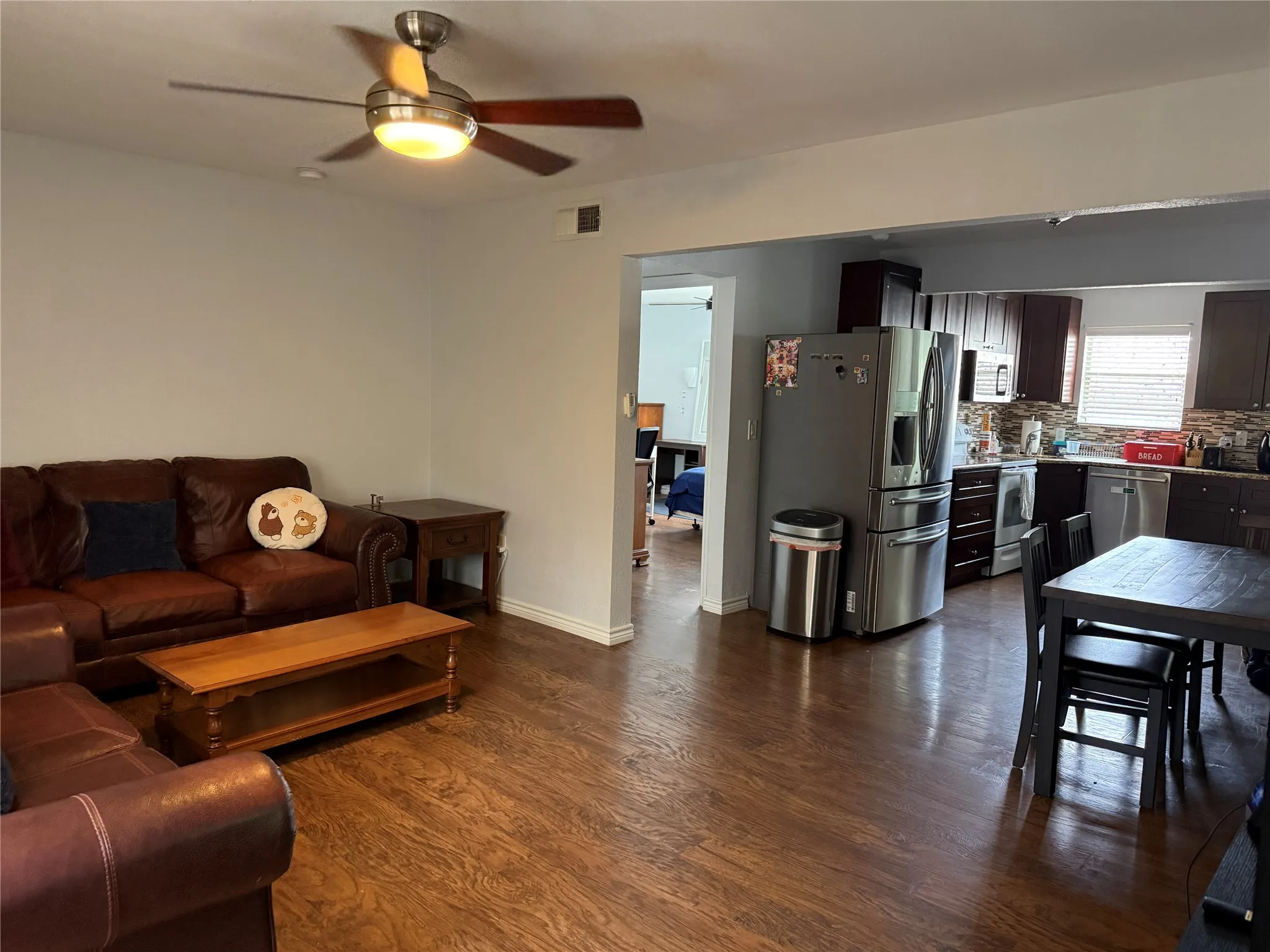 Open-concept living space featuring wood-finish flooring, a ceiling fan with integrated lighting, and a kitchen area with dark cabinetry, stainless steel appliances, and a tiled backsplash