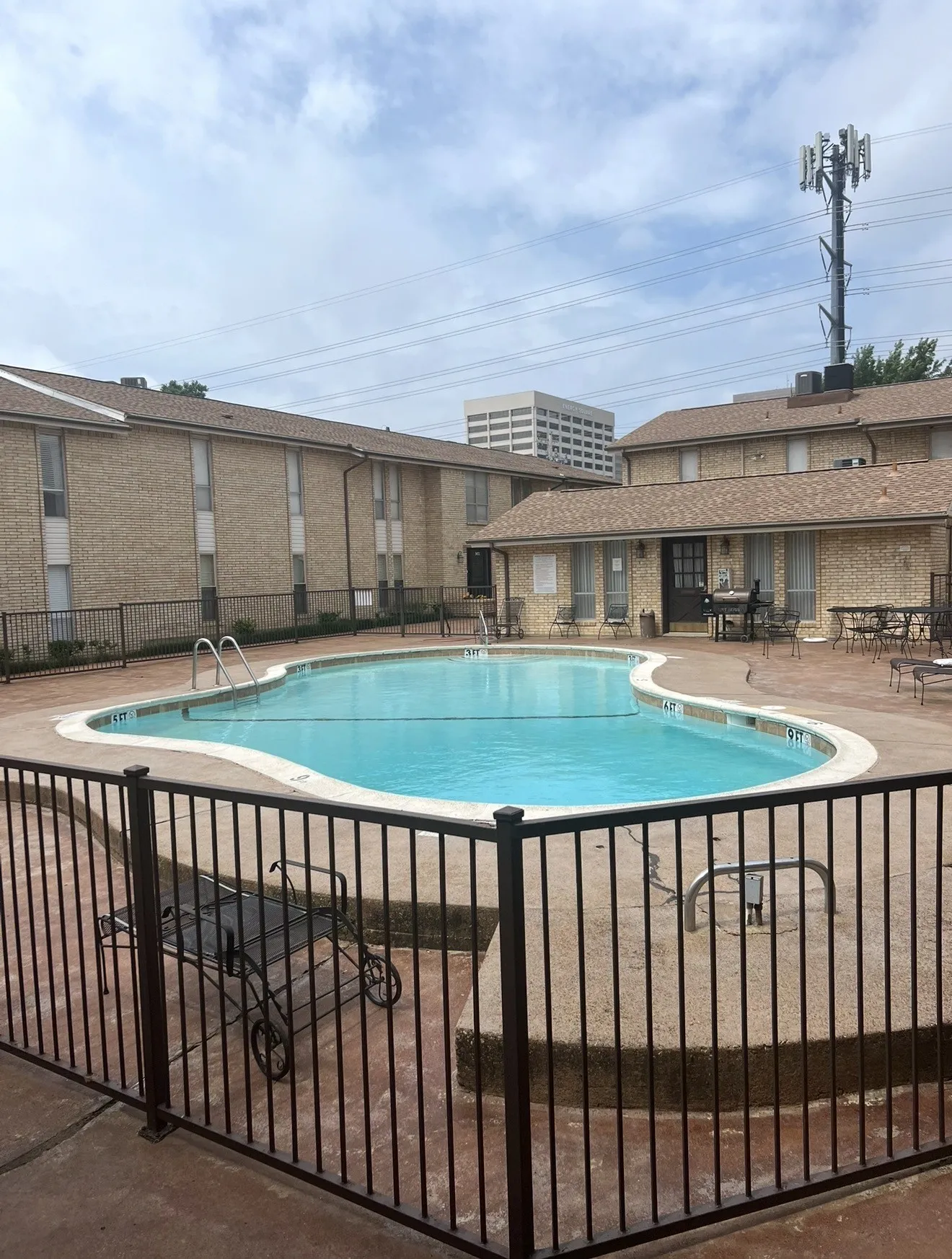 Outdoor pool with surrounding perimeter fencing, concrete decking, and brick-clad buildings