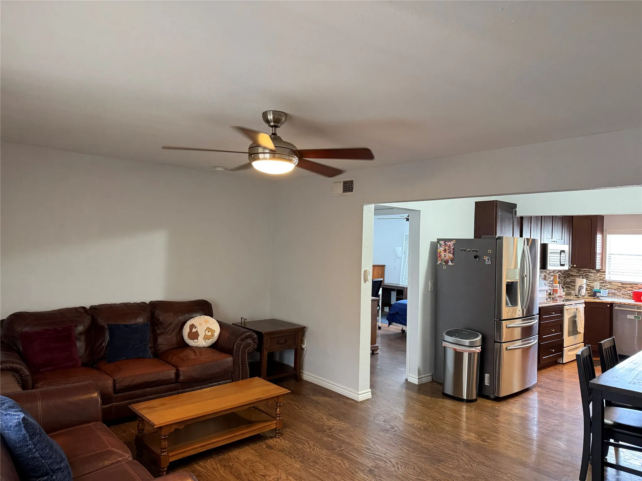 Open-concept living area featuring wood-finish flooring, a ceiling fan with integrated lighting, and a wide entryway into the kitchen