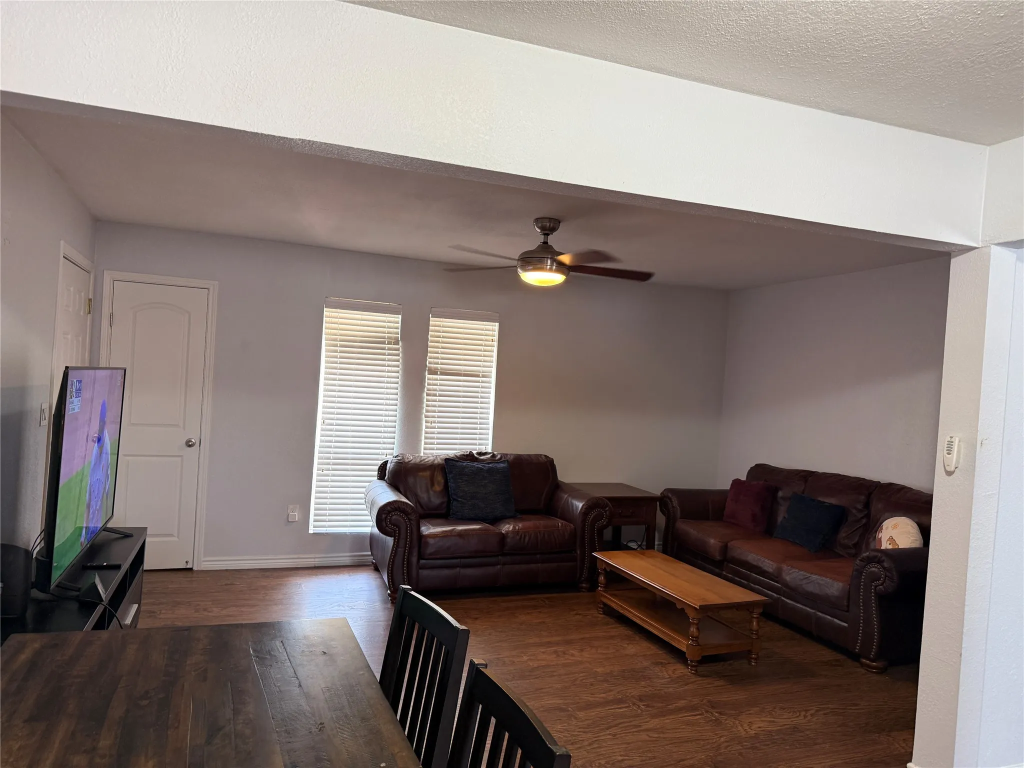 Open-concept living space featuring wood-finish flooring, a ceiling fan with integrated lighting, and two windows with horizontal blinds