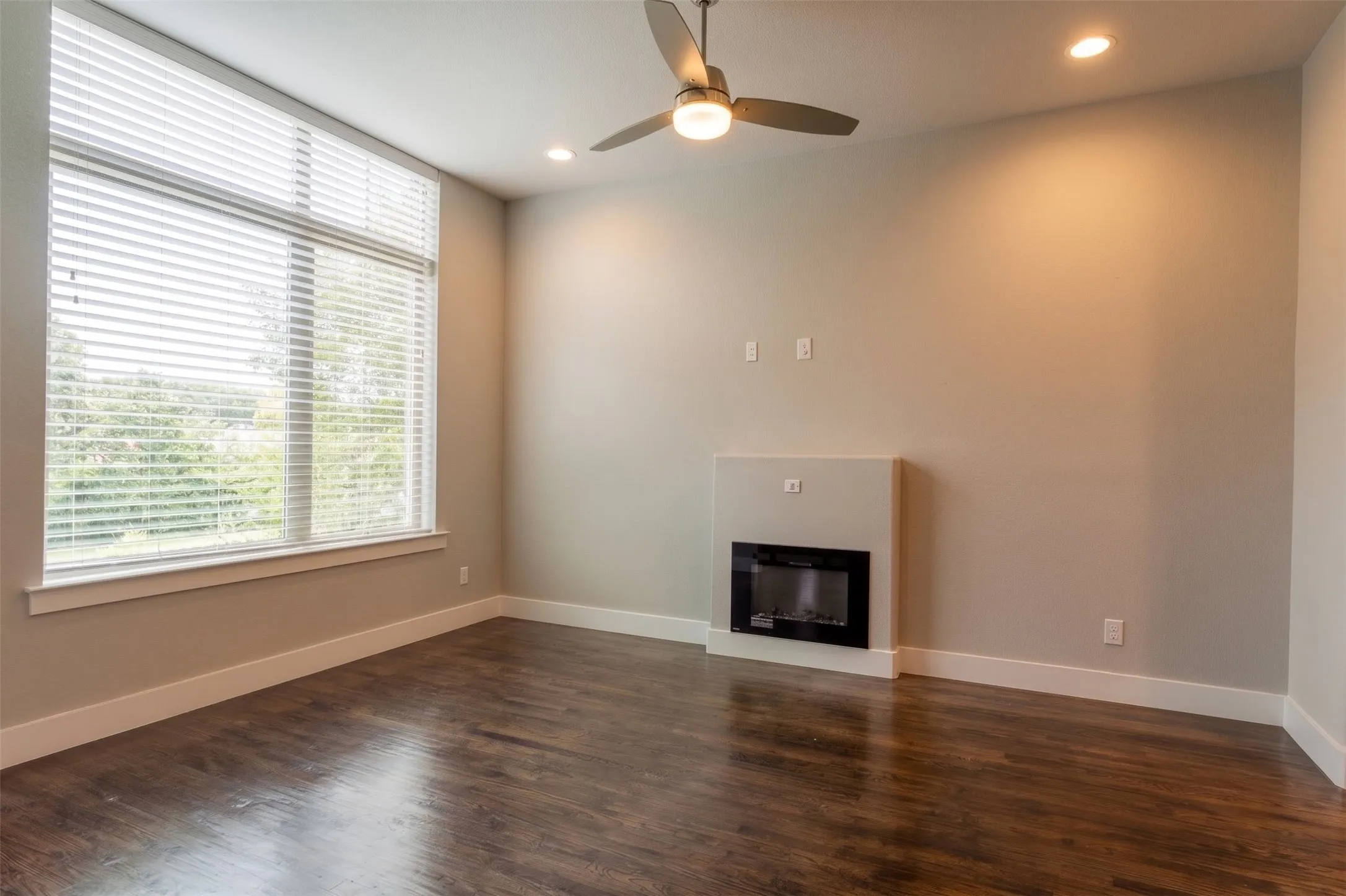 Spacious living room featuring wood-finish flooring, a contemporary built-in electric fireplace, and expansive windows fitted with blinds