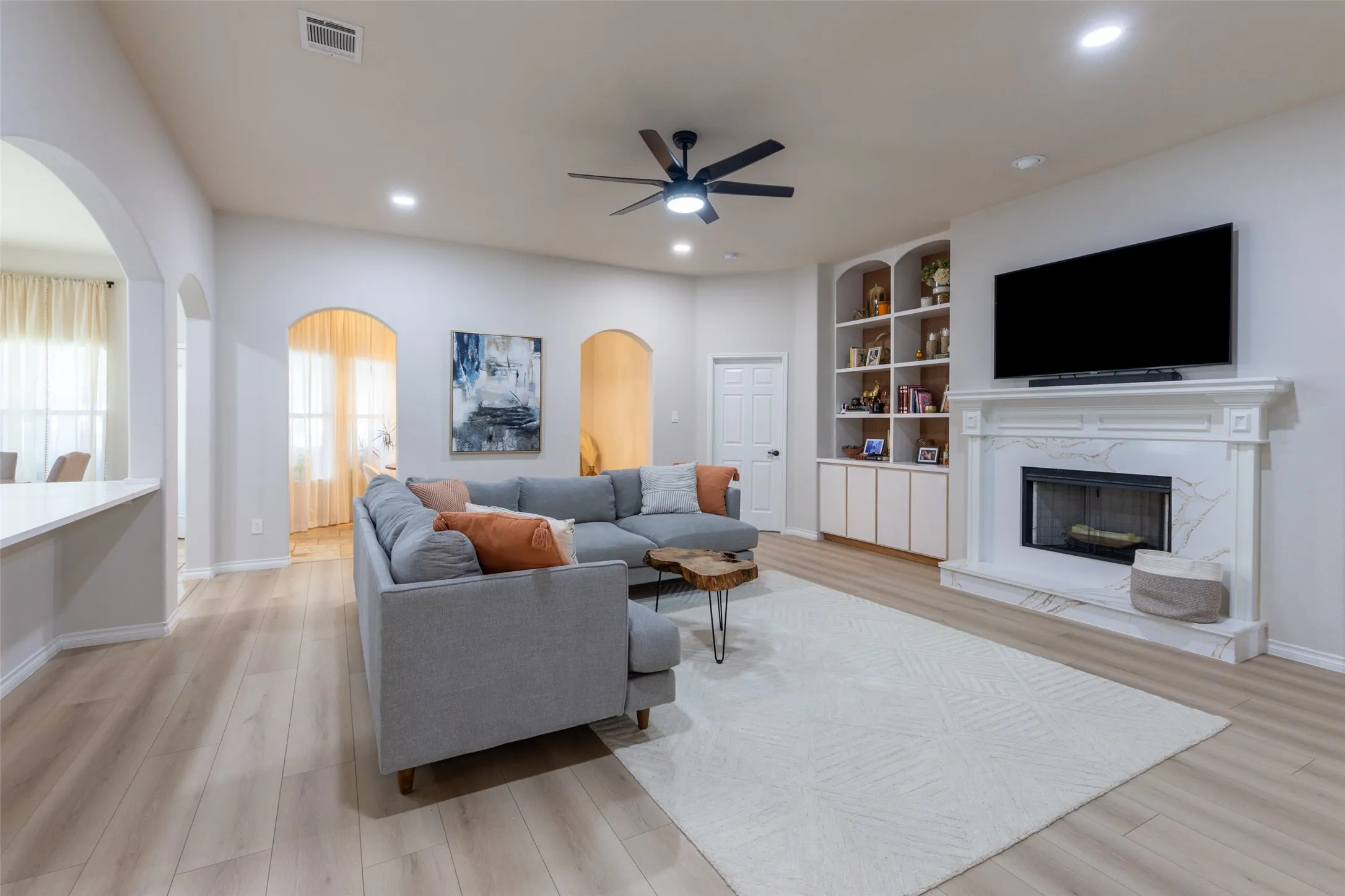 Spacious living area featuring wood-finish flooring, a ceiling fan, recessed lighting, and a white fireplace with a mantel