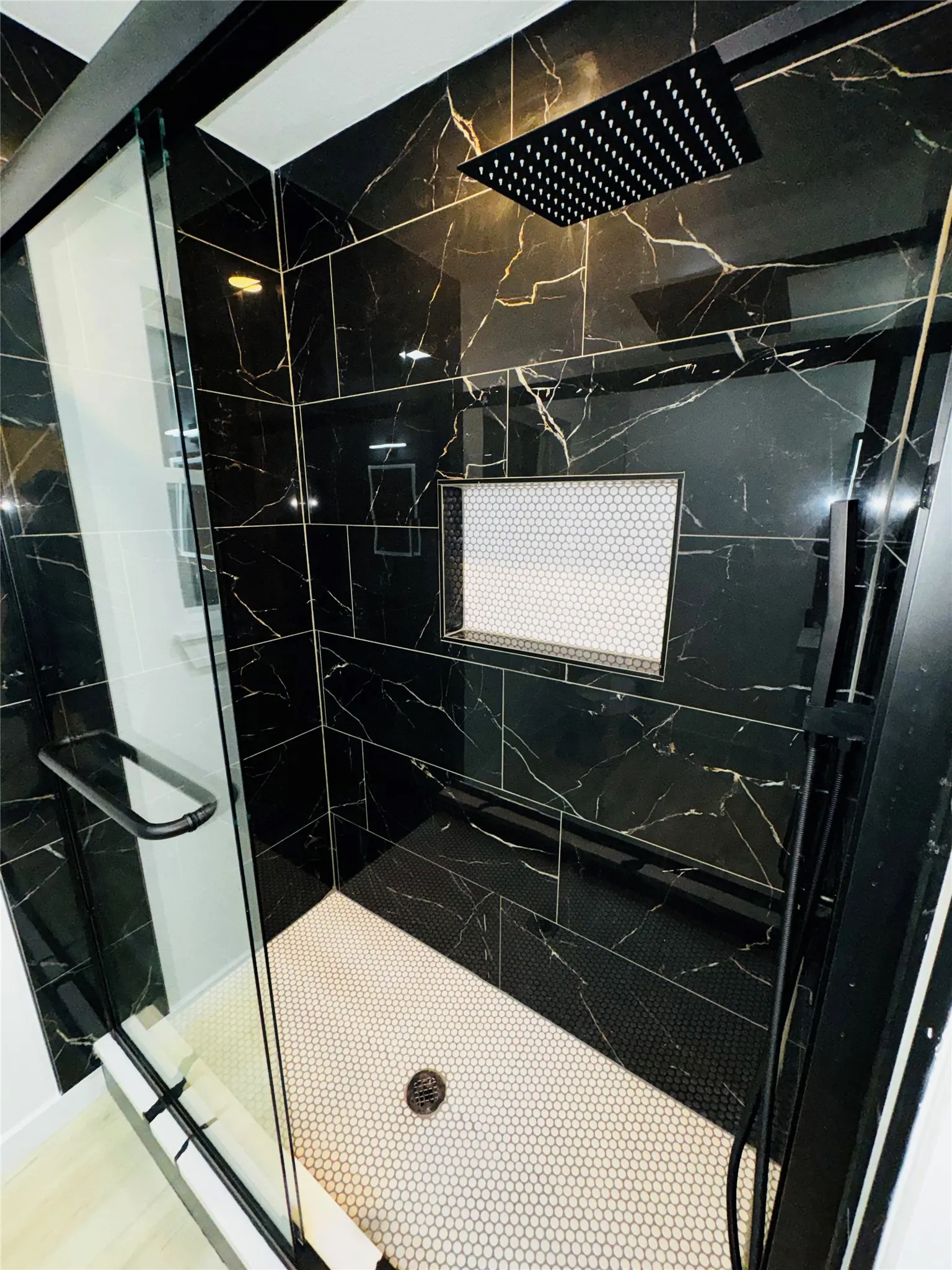 Black marble-look tile shower enclosure with a rainfall shower head, glass shower door, built-in tiled niche, and white hex tile flooring