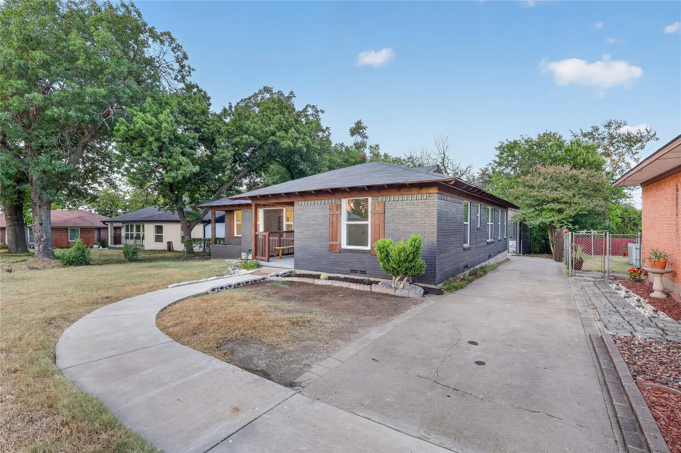 Exterior featuring dark brick siding, decorative wooden shutters, a covered front porch, a paved driveway, and mature trees