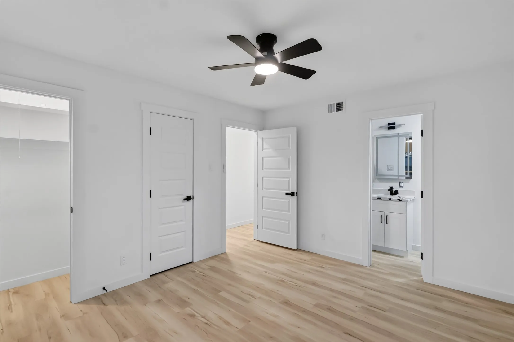 Light wood-finish flooring, a modern black ceiling fan with integrated lighting, flat panel interior doors, and white painted walls