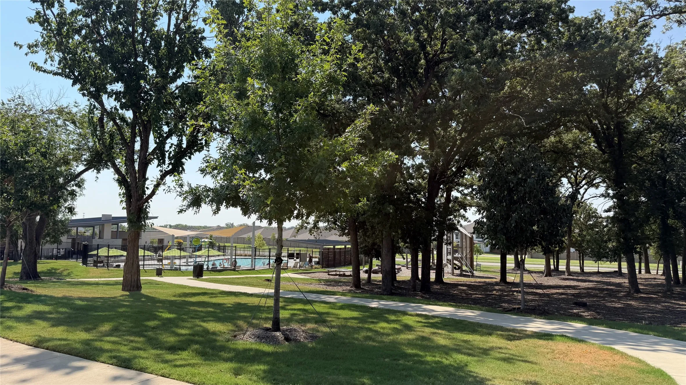 Expansive green space featuring mature trees, a paved pathway, and a distant recreational area with a swimming pool and covered lounge structures