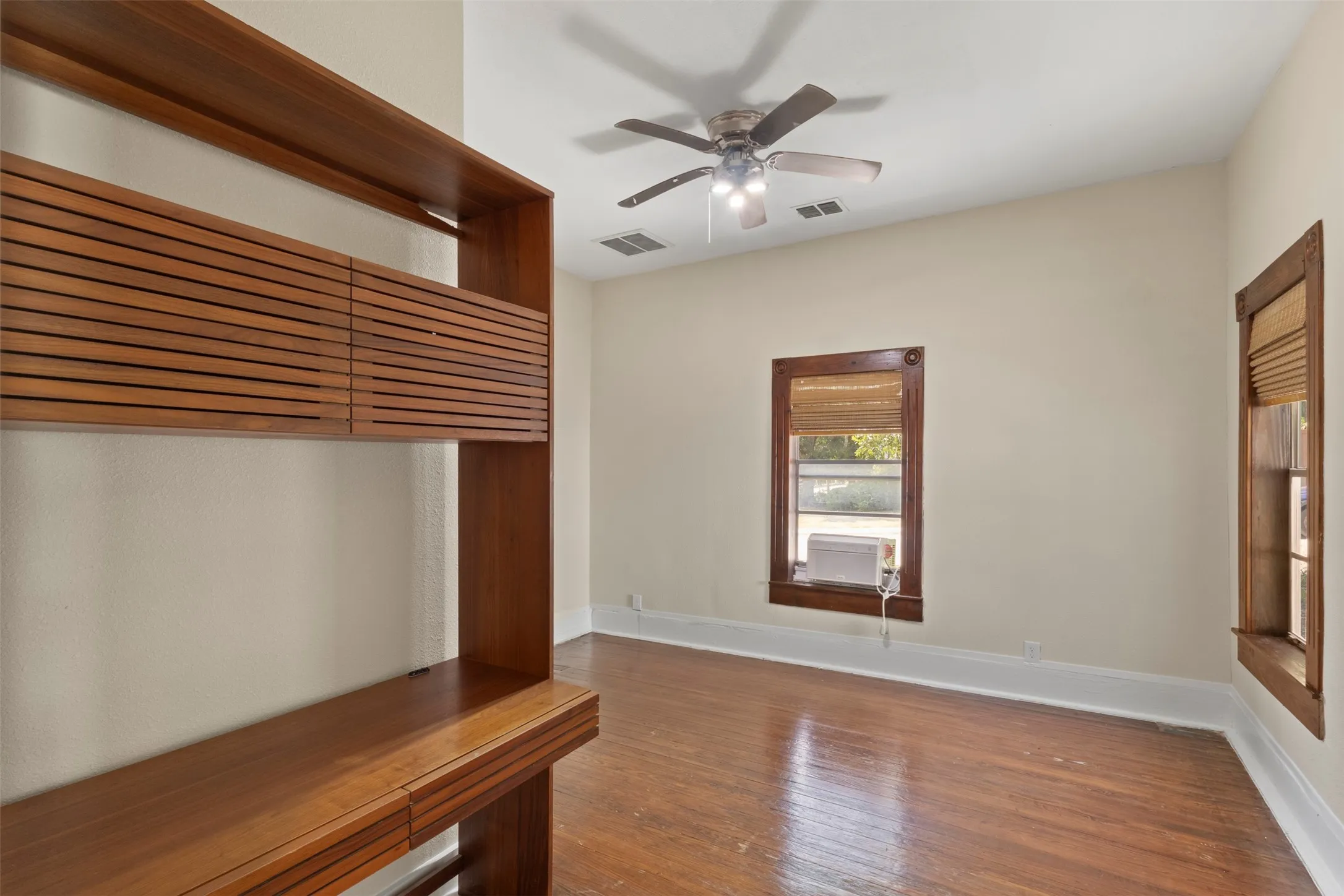 Built-in wood-finish shelving and desk unit, wood-finish flooring, two large windows, and a ceiling fan with integrated lighting