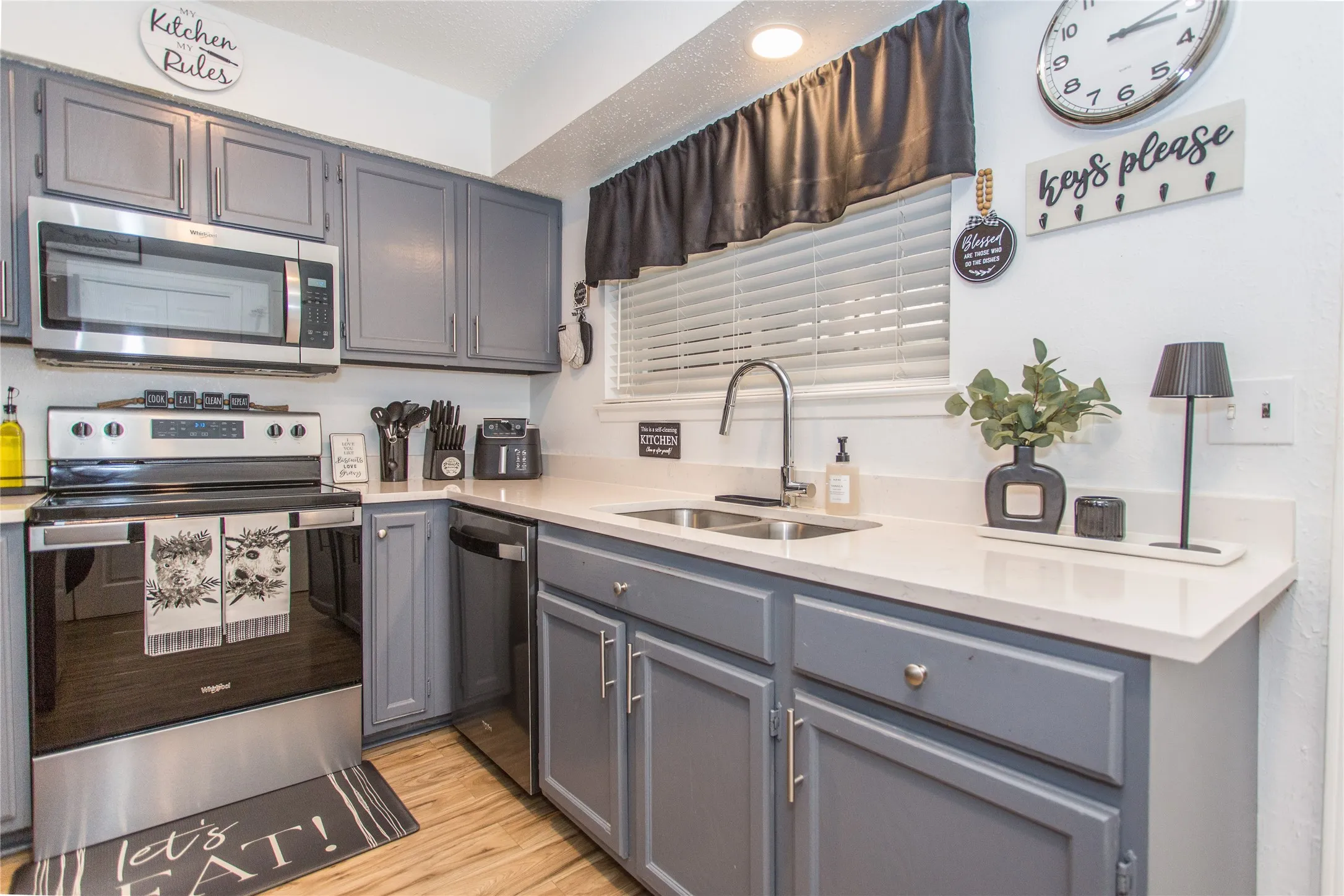 Kitchen featuring grey cabinetry with silver hardware, stainless steel appliances, a double basin sink with a gooseneck faucet, light-toned countertops, and wood-finish flooring