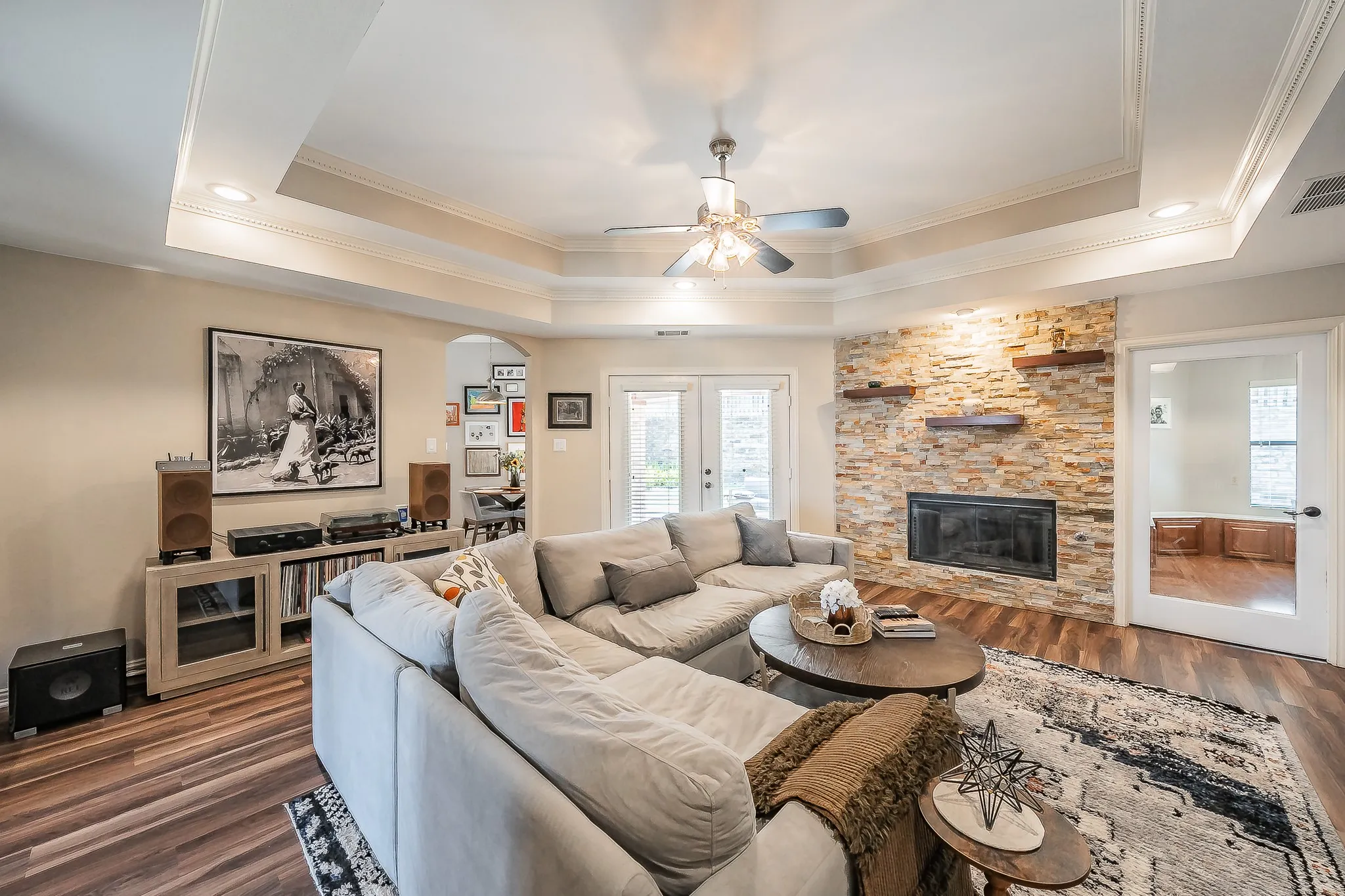 Living area featuring tray ceilings with crown molding, recessed lighting, a ceiling fan, a stone-clad fireplace, and double glass-paneled doors
