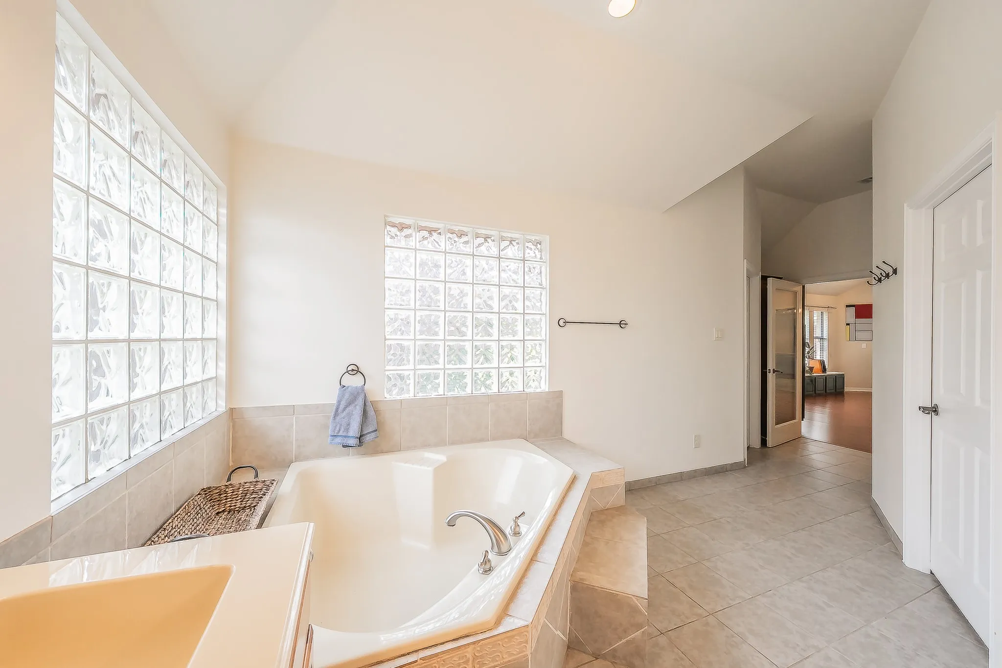 Built-in corner soaking tub with surrounding tile, two large glass block windows, vaulted ceiling, and light-toned tile flooring