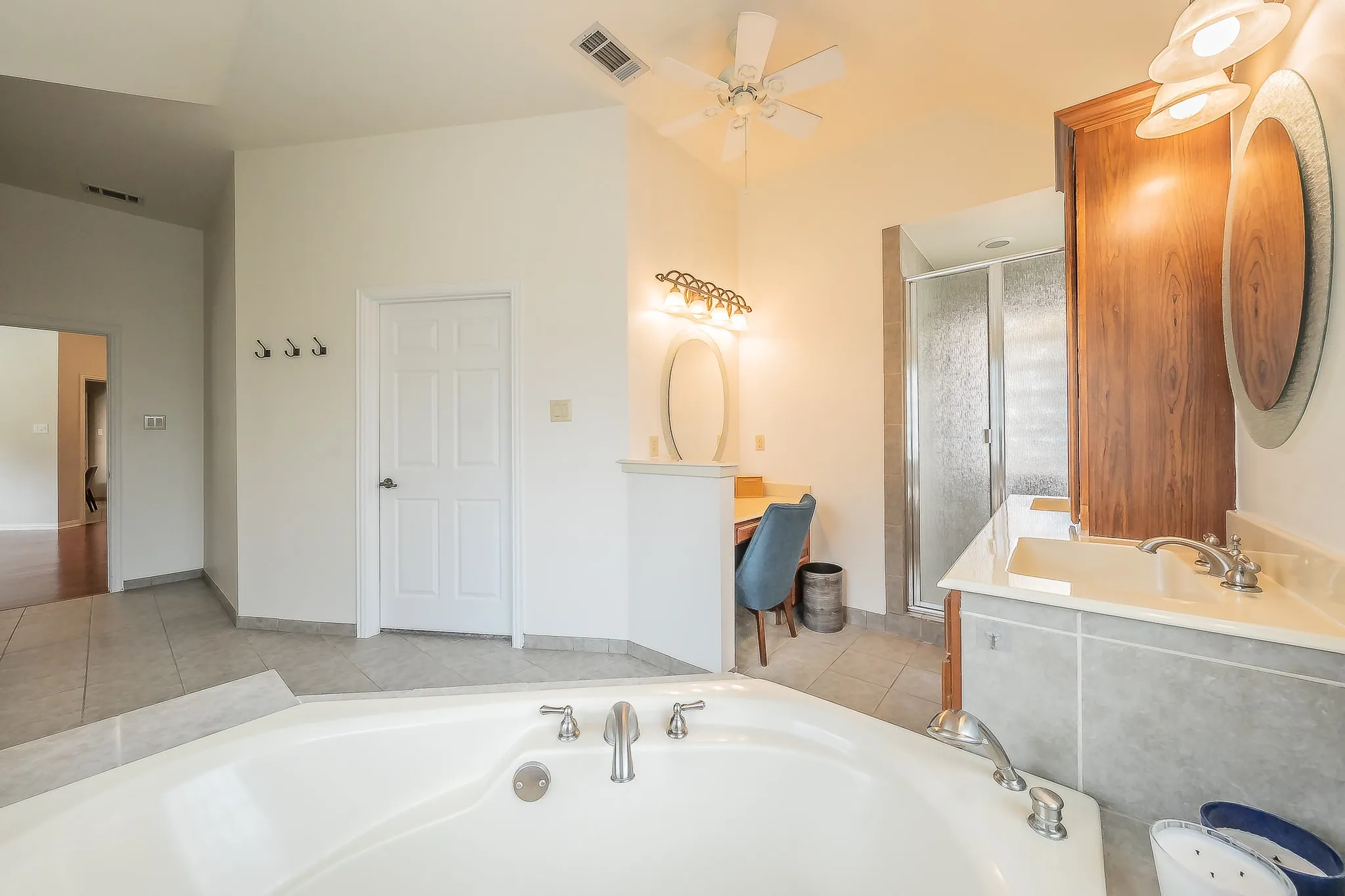 Corner soaking tub with deck-mounted fixtures, separate shower enclosure, dual vanity sinks with light-toned countertops, and ceiling fan