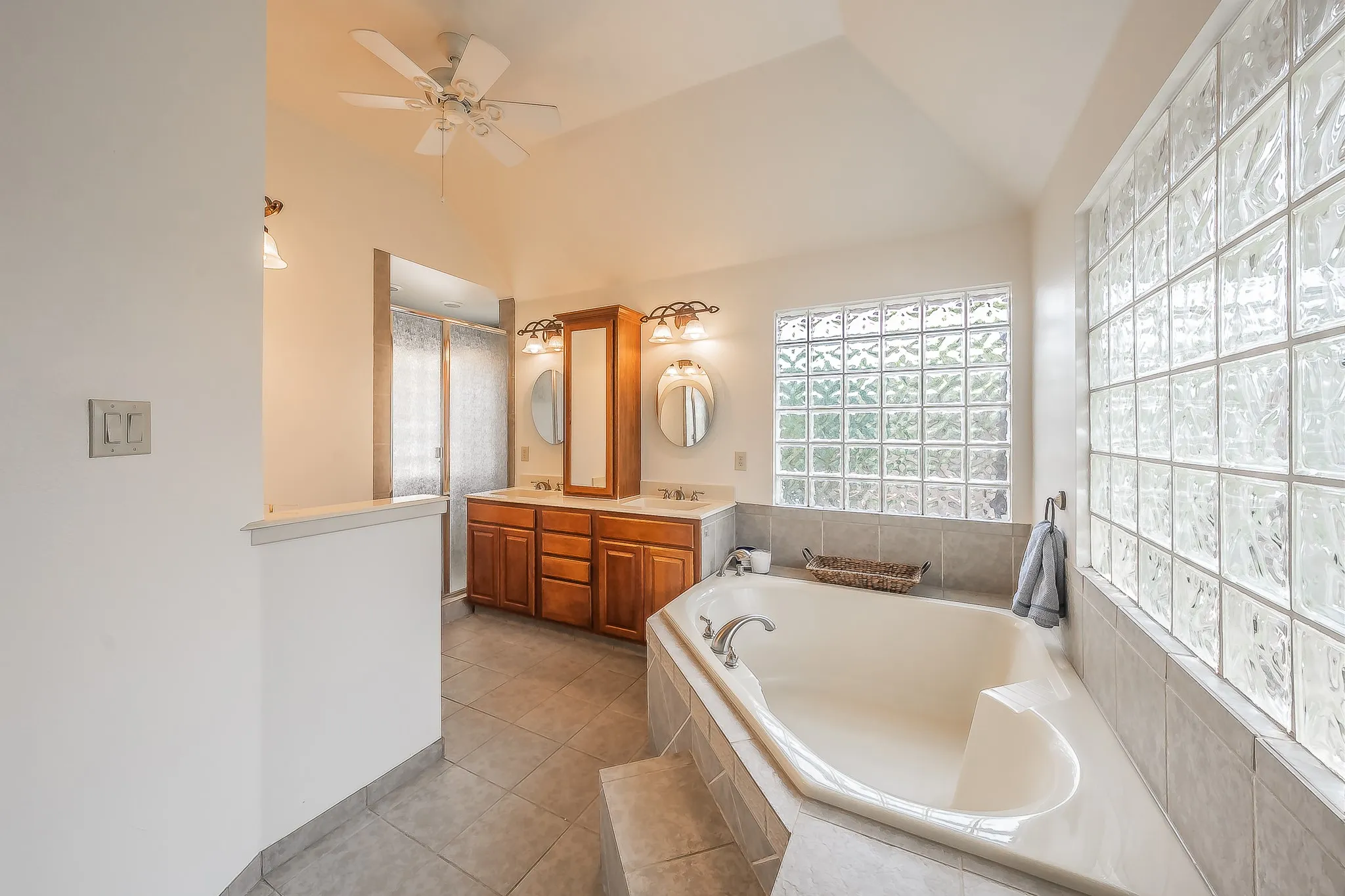 Vaulted ceiling with ceiling fan, dual vanity with wood-finish cabinetry, built-in soaking tub, and block glass windows
