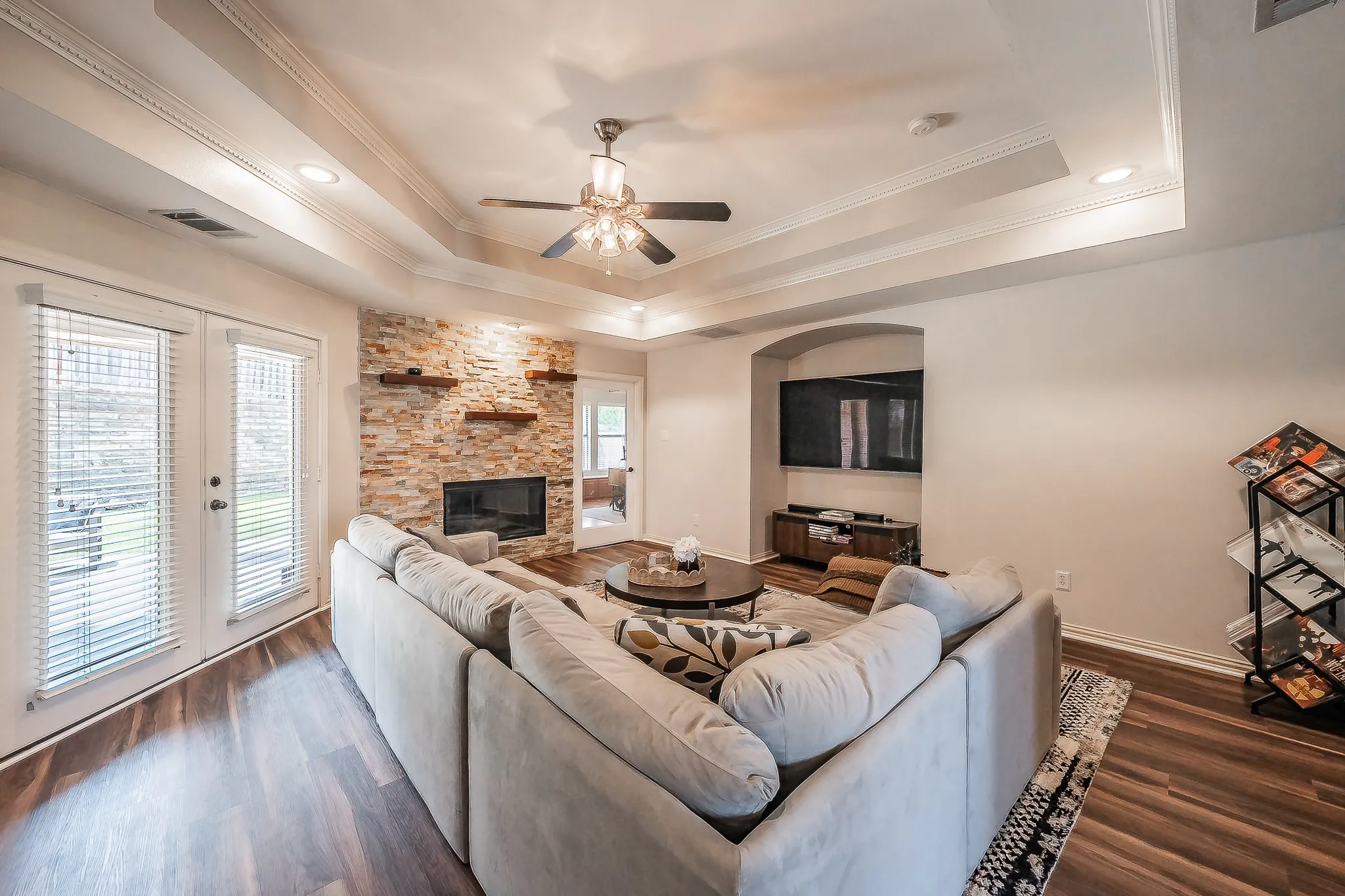 Wood-finish flooring, stone-clad fireplace with hearth, recessed lighting, double tray ceiling, and ceiling fan with integrated lighting