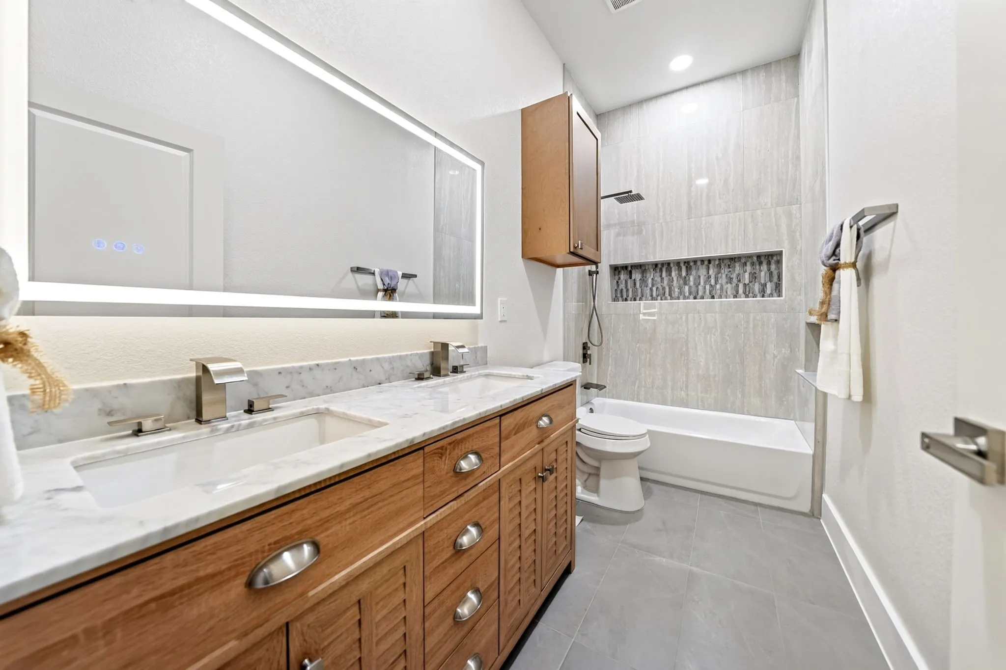 Dual vanity with integrated sinks and marble-look countertop, large back-lit mirror, built-in bathtub with tiled surround, and a recessed shower niche with mosaic tile