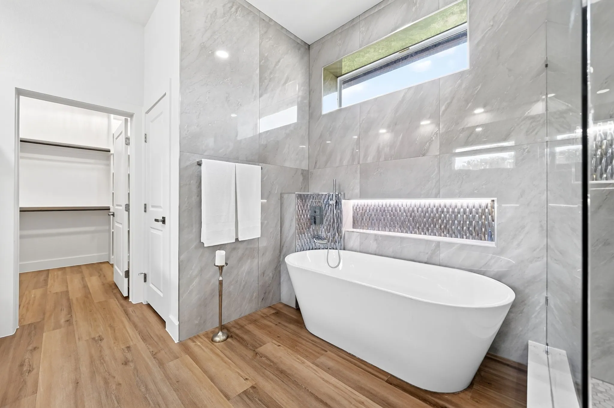 Freestanding soaking tub with a wall-mounted faucet, complemented by a tiled accent niche, wood-finish flooring, and a walk-in closet with built-in shelving