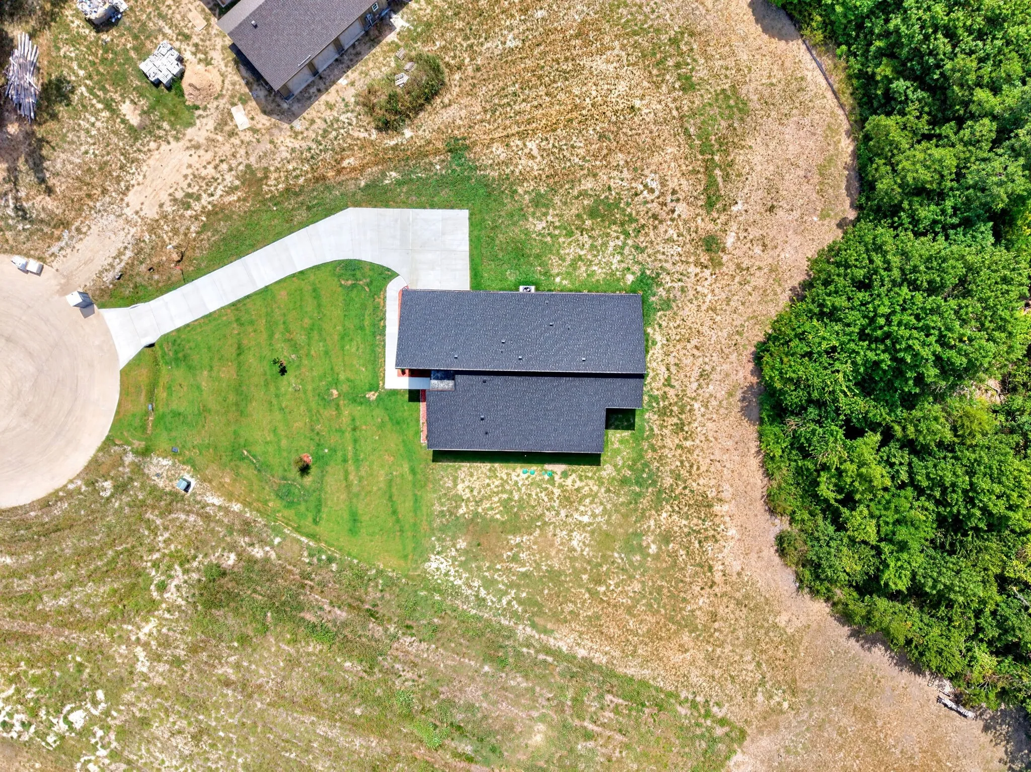 Newly constructed residence with a dark shingle roof, concrete driveway, and a partially landscaped yard, bordered by mature trees