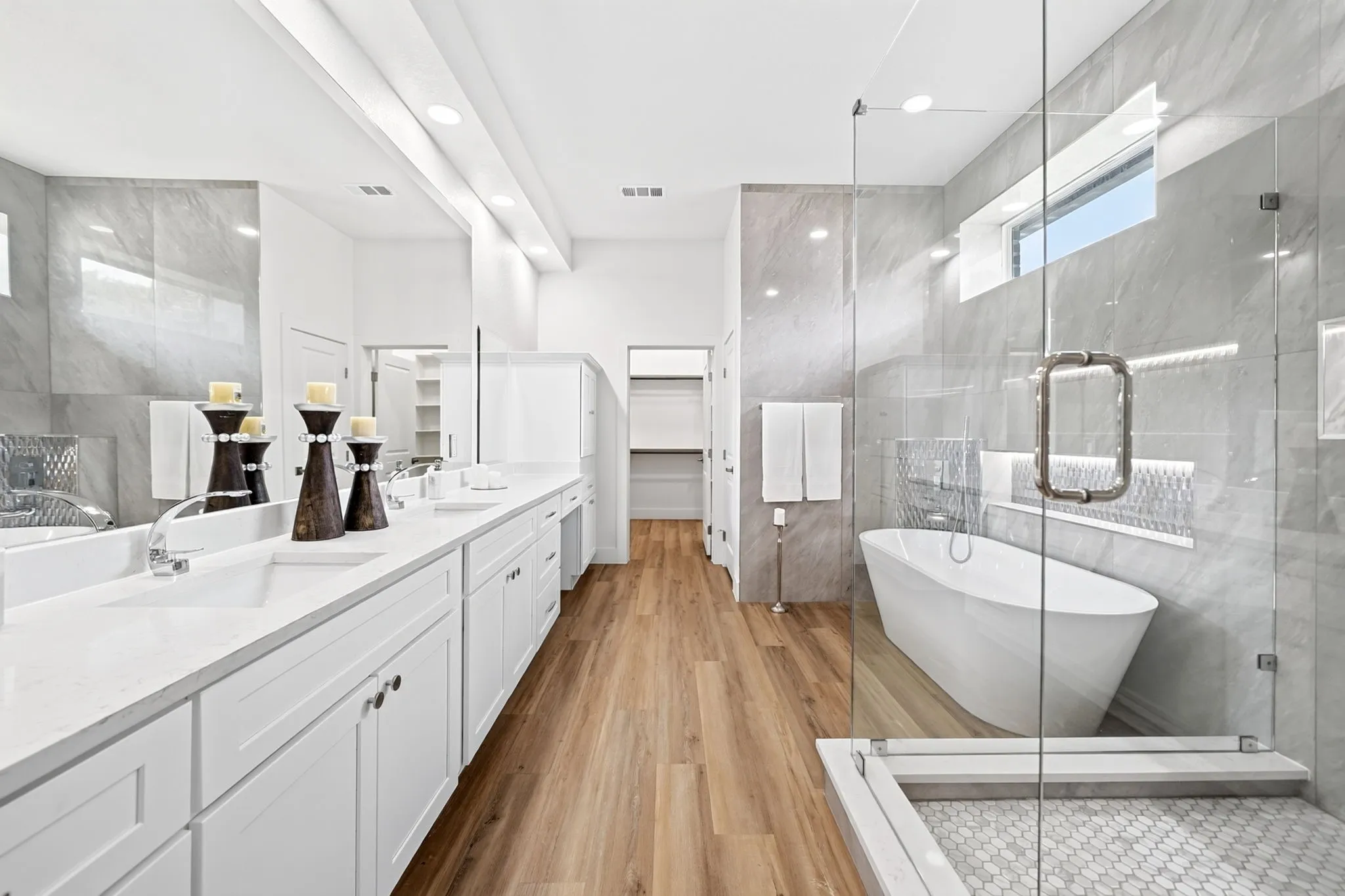 Dual vanity with white cabinetry and integrated sinks, wood-finish flooring, freestanding soaking tub, and a glass-enclosed shower with tiled walls