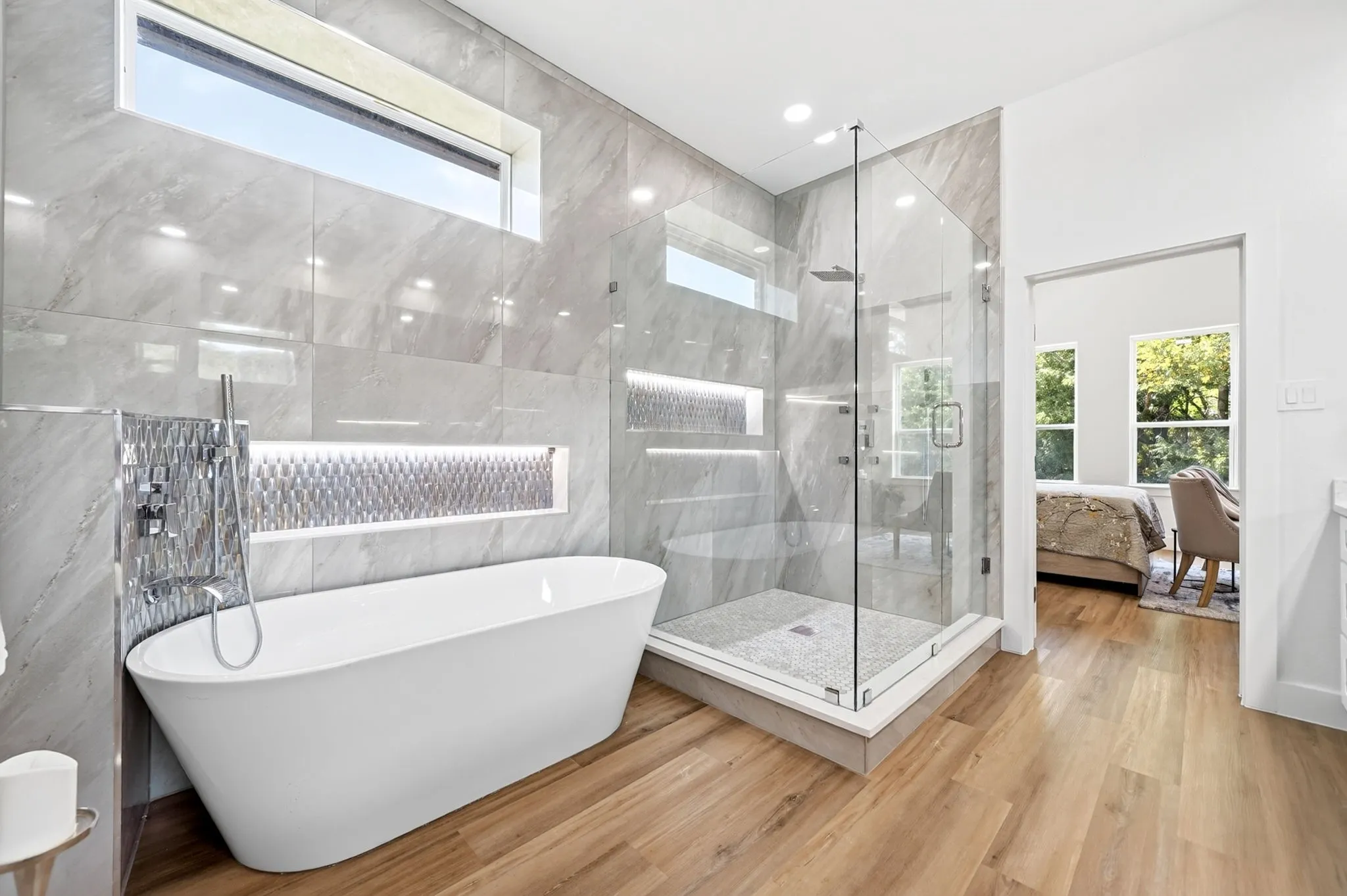 Freestanding soaking tub, glass-enclosed shower, wood-finish flooring, tiled walls with recessed lighting, and transom window