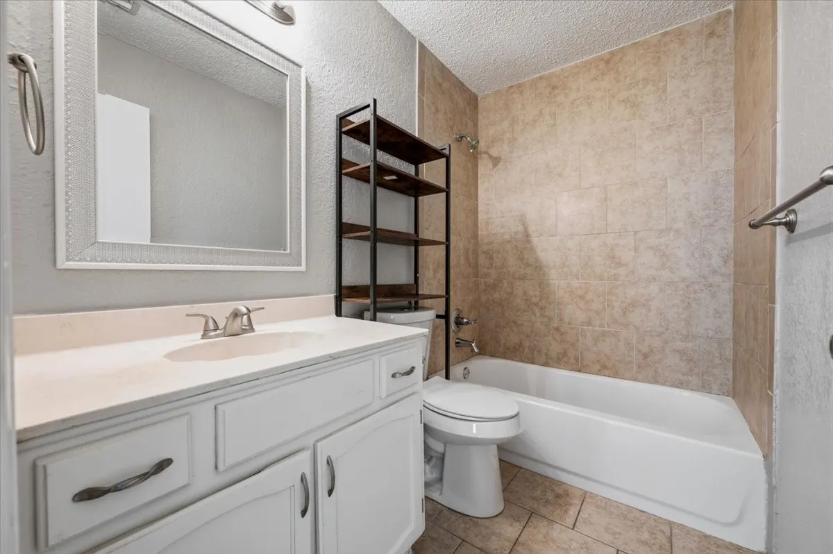 Primary bathroom features a vanity with mirror, and a tub/shower combo with full tile surround. A built-in shelving unit sits above the toilet for storage. Tile flooring extends throughout.