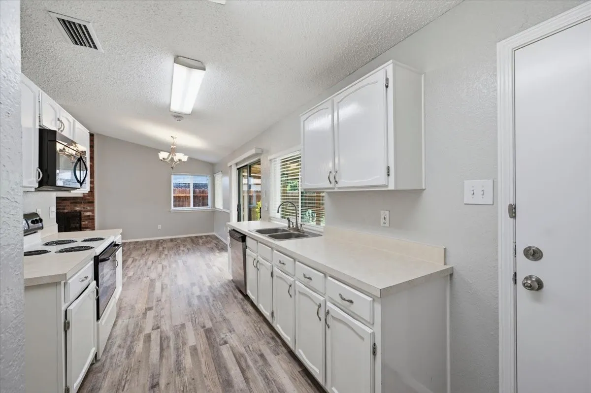 Kitchen features white cabinetry, a stainless dishwasher, and a sliding glass door leading to the covered patio. A window over the sink and the connected dining area with chandelier are visible beyond. Wood-look vinyl flooring extends throughout.