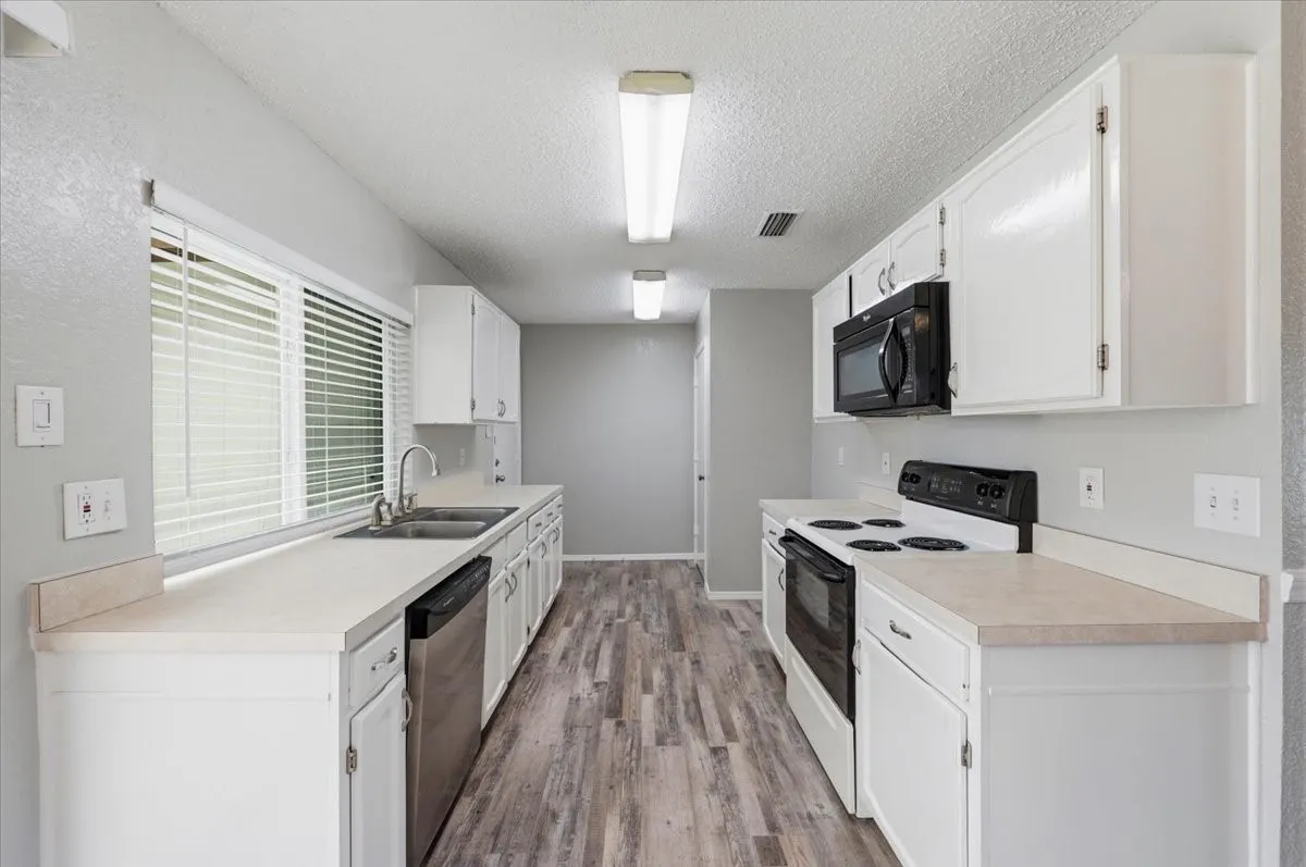 Kitchen features white cabinetry with a built-in microwave, and a stainless dishwasher. A window over the sink brings in natural light. The layout connects to the pantry and garage entry beyond. Wood-look vinyl flooring extends throughout.