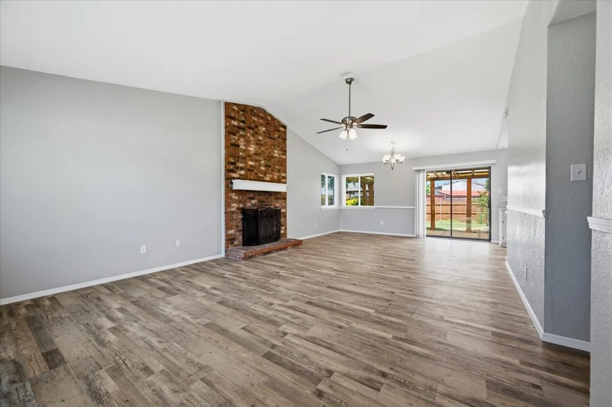 Living room features a vaulted ceiling with ceiling fan, and a brick fireplace extending floor to ceiling. A sliding glass door leads to the covered patio, with windows above the adjoining dining space. The room opens directly into the dining area. Wood-look vinyl flooring extends throughout.