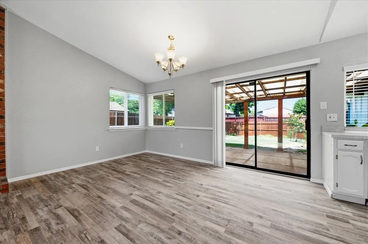 Dining area features a vaulted ceiling with light fixture. Large windows overlook the back yard, and a sliding glass door opens to the covered back patio. The space connects directly to the adjoining kitchen. Wood-look vinyl flooring extends throughout.