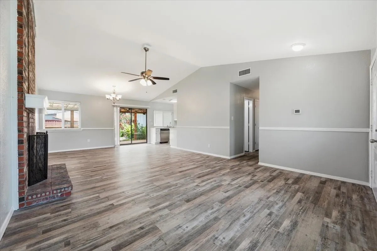 Living room features a vaulted ceiling with ceiling fan, and a brick fireplace hearth. A window over the kitchen sink is visible beyond, with the layout opening to both the kitchen and hallway. Wood-look vinyl flooring extends throughout, transitioning toward the kitchen.