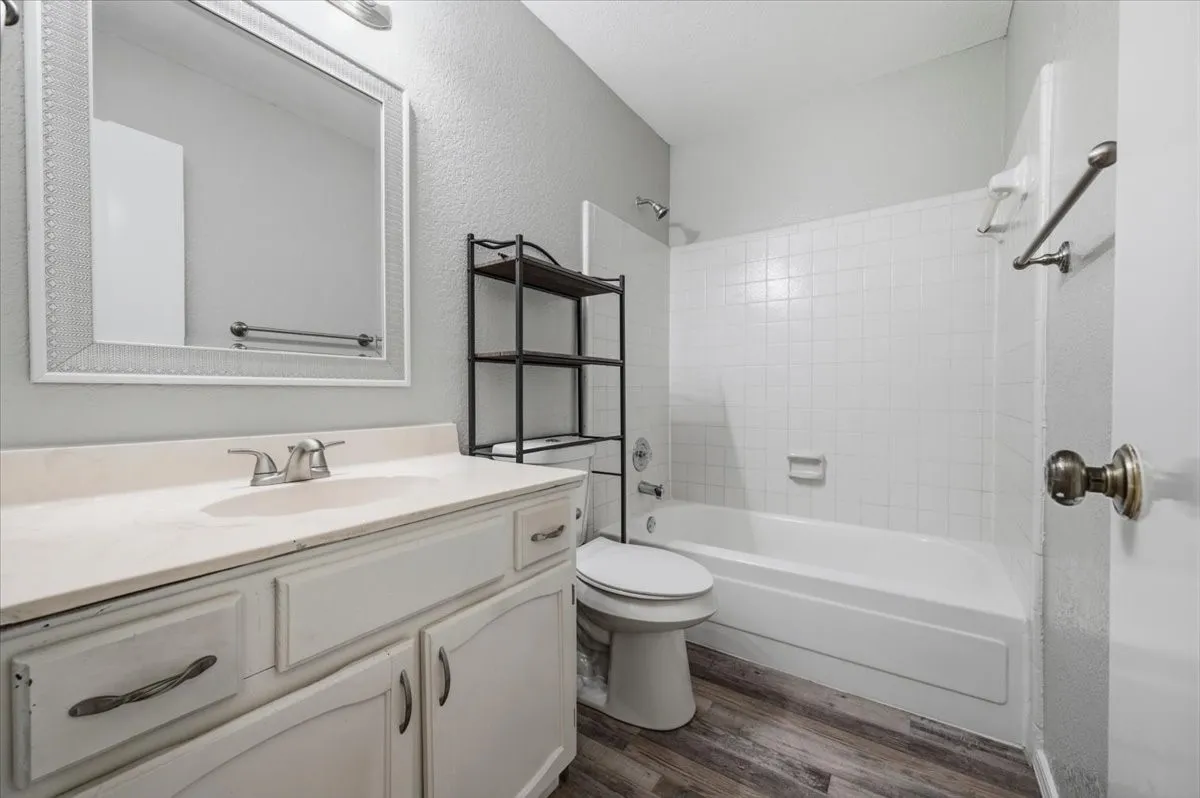 Secondary bathroom features a vanity with mirror, and a tub/shower combo with tile surround. A built-in shelving unit sits above the toilet for additional storage. Wood-look vinyl flooring extends throughout.