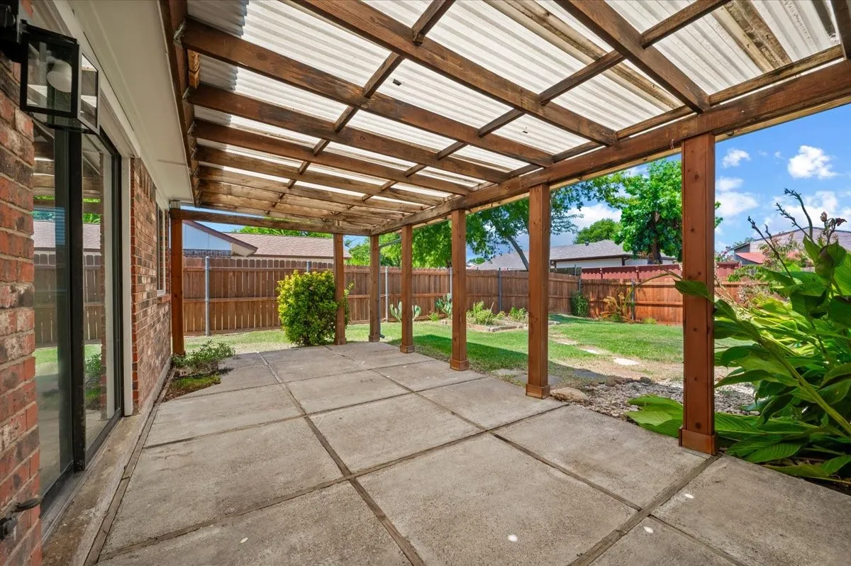 Covered patio features a wood pergola-style structure with corrugated polycarbonate roofing. A sliding glass door connects to the interior, and the space overlooks a privacy-fenced backyard with mature landscaping.