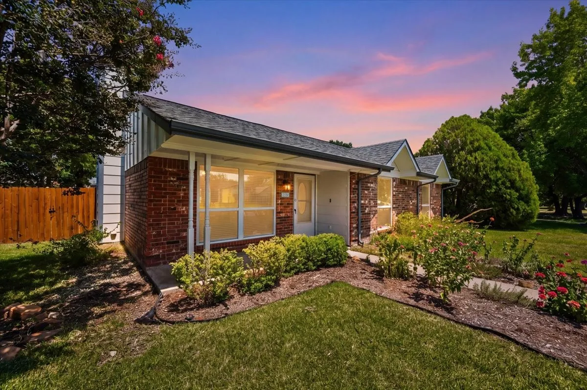 Front exterior features a covered porch with painted wood columns and brick veneer siding. Large windows flank a paneled front door with glass sidelight. A concrete walkway winds through landscaped flower beds, with a wood privacy fence enclosing the side yard.
