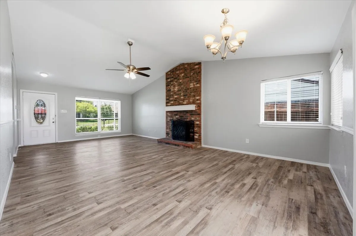 Living room features a vaulted ceiling with ceiling fan and light fixture, plus a brick fireplace with wood mantel. A front door with glass sidelight and large window bring in natural light. The space opens to the dining area, visible in the background. Wood-look vinyl flooring extends throughout.