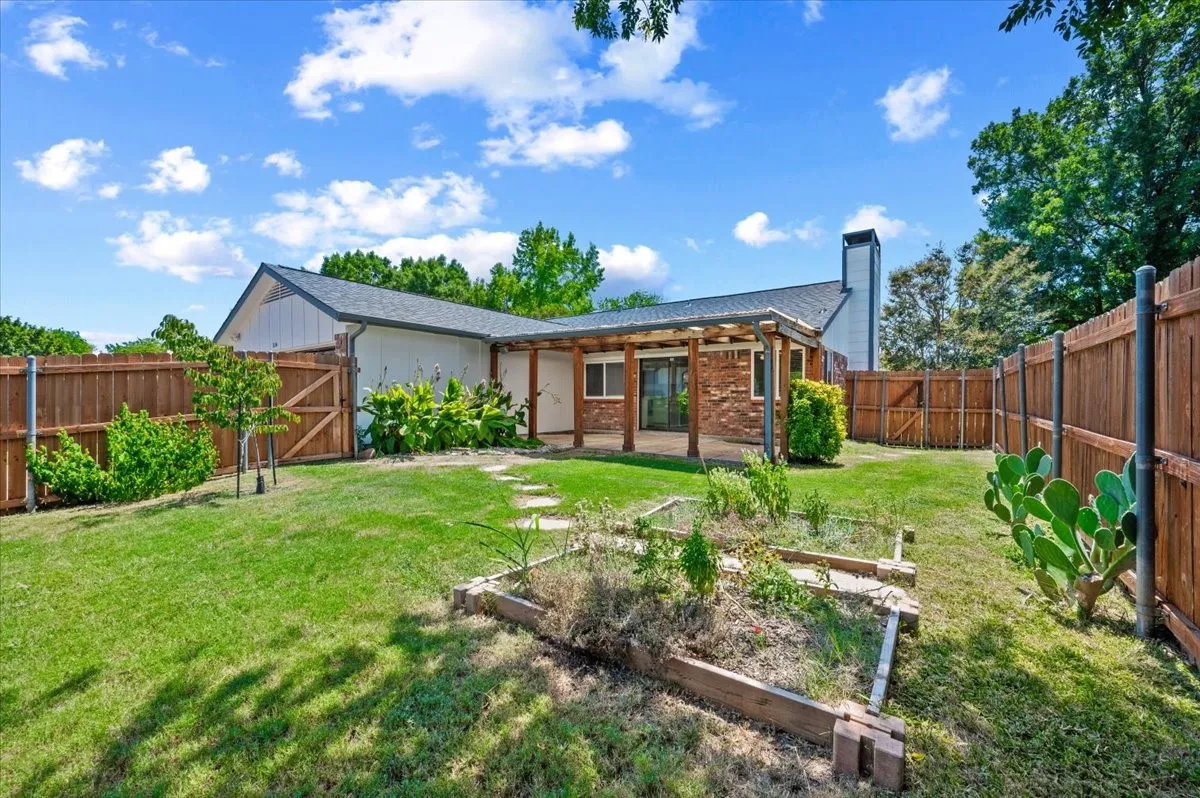 Backyard features a covered patio area with wood pergola structure, surrounded by privacy fencing. Raised garden beds and a stepping-stone path lead through the mature landscaping, with a brick chimney visible on the rear of the home.