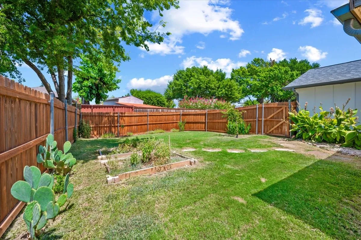 Backyard features privacy fencing enclosing a grassy yard with raised garden beds and mature trees. A side gate provides access, with additional landscaping along the fence line.