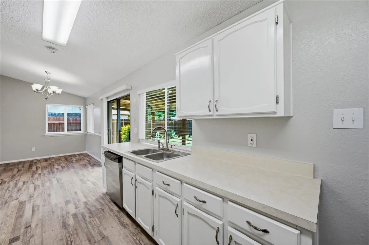 Kitchen features white cabinetry above a stainless dishwasher, with a sliding glass door and window over the sink bringing in natural light. The layout opens to the adjoining dining area. Wood-look vinyl flooring extends throughout.
