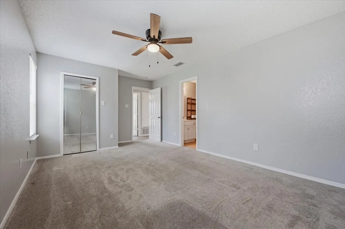 Primary bedroom features a ceiling fan with light fixture and a mirrored sliding closet door. The layout opens to the hallway and to the ensuite bath, with cabinetry visible beyond. Carpet flooring extends throughout.